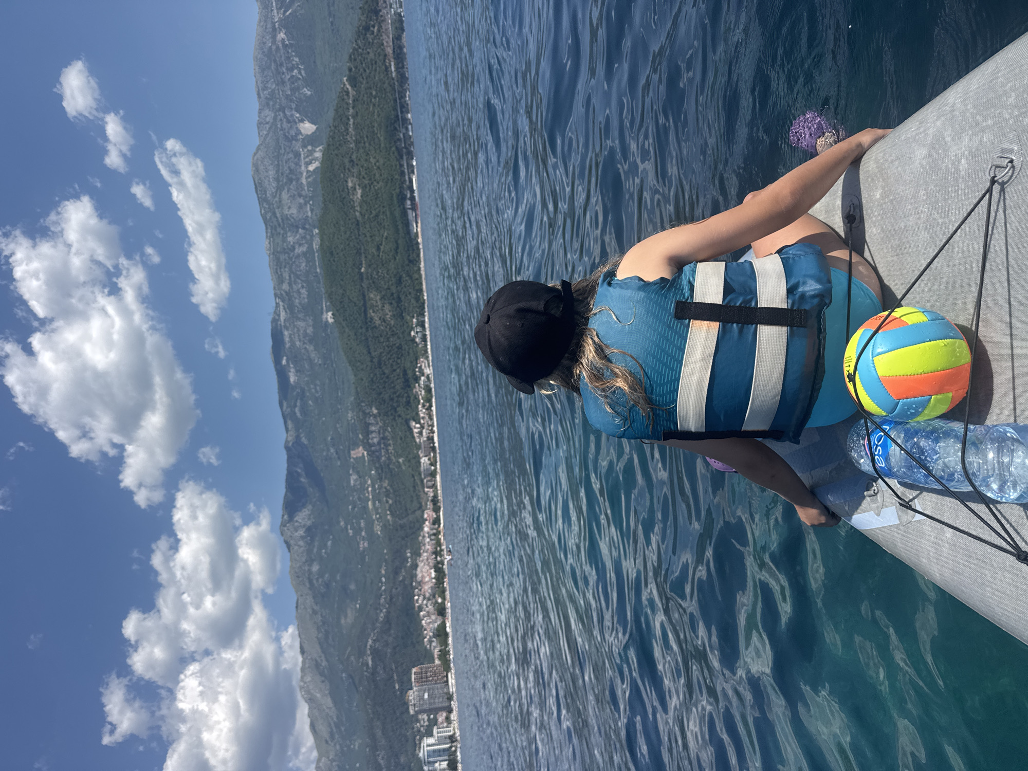 A person wearing a black cap and a blue and white life vest is sitting on a paddleboard in the Adriatic Sea, facing away from the camera. The person is wearing blue swim bottoms. A colorful ball and a bottle of water are on the paddleboard in front of the person. In the background, the city of Budva, Montenegro, with its buildings and hotels, is visible along the coastline. Majestic mountains rise behind the city, and the sky is blue with scattered white clouds. The water is a clear, deep blue with gentle ripples.