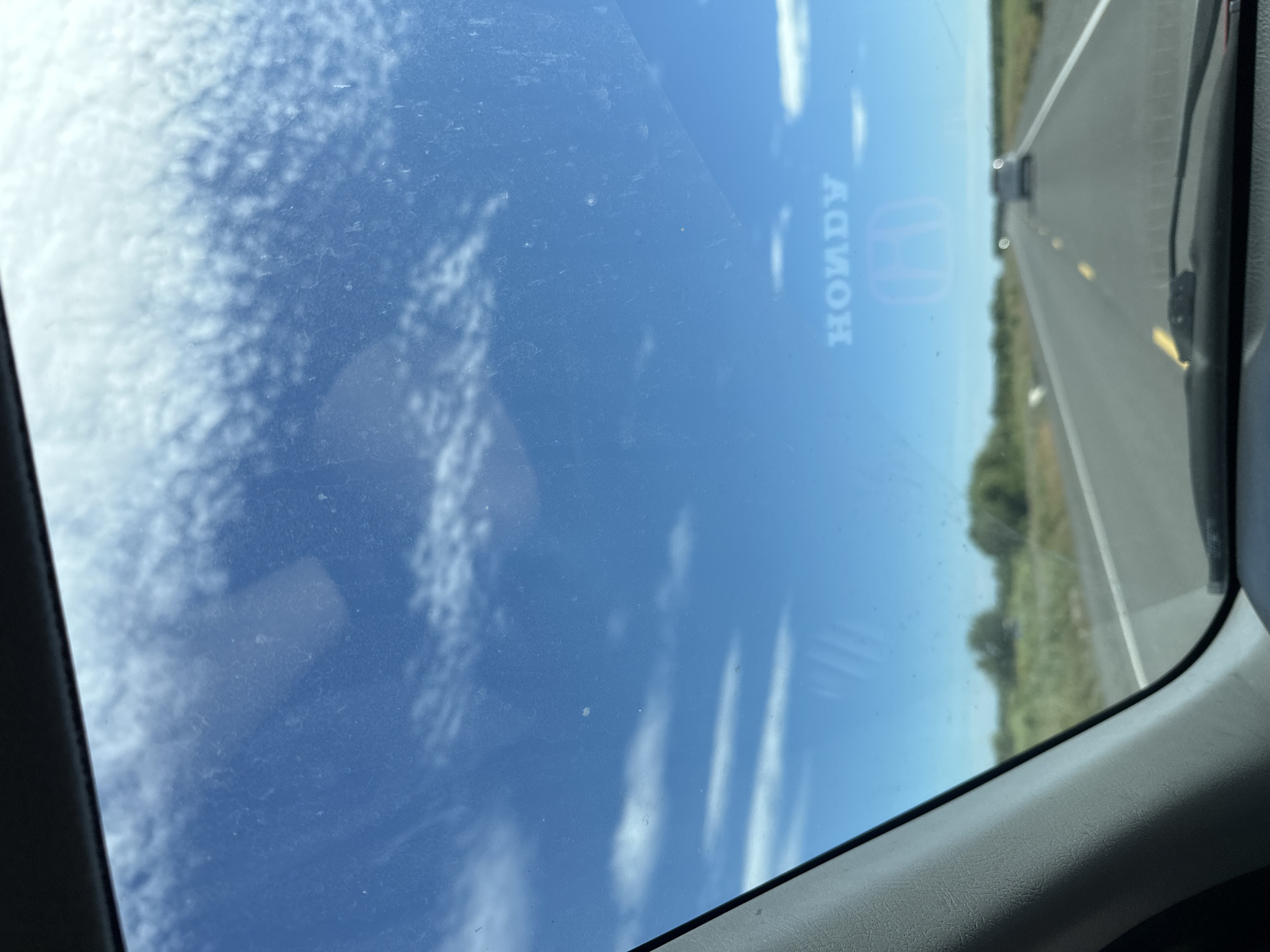 The image shows a view from inside a vehicle looking out through the windshield. The sky is blue with some scattered clouds. A highway stretches into the distance, with trees and vegetation on either side. There are a few other vehicles on the road ahead. A faint reflection of the "HONDA" logo and the Honda emblem is visible on the windshield. The scene appears to be a rural or suburban area. There is no information in the image to indicate the location is Petropavlovsk-Kamchatsky, Russia.