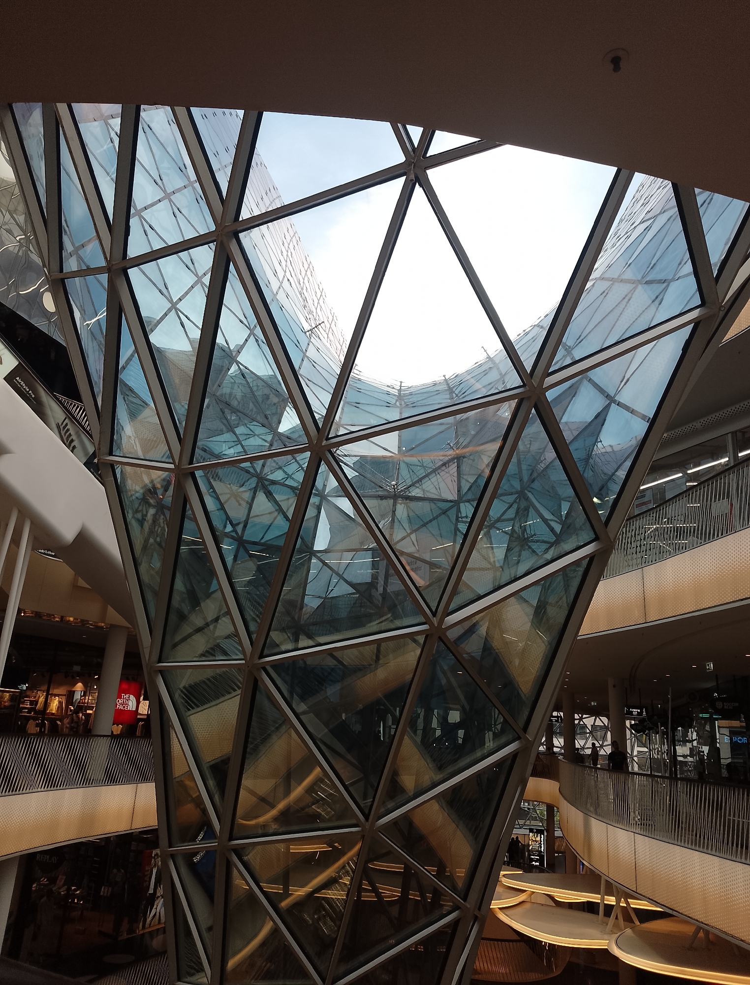 The image depicts the interior of a modern, multi-story building with a striking geodesic dome-like glass structure at its center. The dome is composed of numerous triangular glass panels framed by dark metal struts, creating a complex geometric pattern. Reflections of the interior and the sky are visible on the glass surfaces.
Multiple levels of the building are visible, characterized by balconies with railings and shops or commercial spaces. Some of these spaces have signage, including one that appears to be "THE NORTH FACE." The lighting suggests it is daytime, with natural light filtering through the glass structure.
Several individuals can be seen on the different levels, some browsing in shops and others walking along the walkways. The overall aesthetic is contemporary and architectural, emphasizing light, reflection, and geometric design. The context appears to be a commercial or retail environment, likely a shopping mall or a public atrium space.