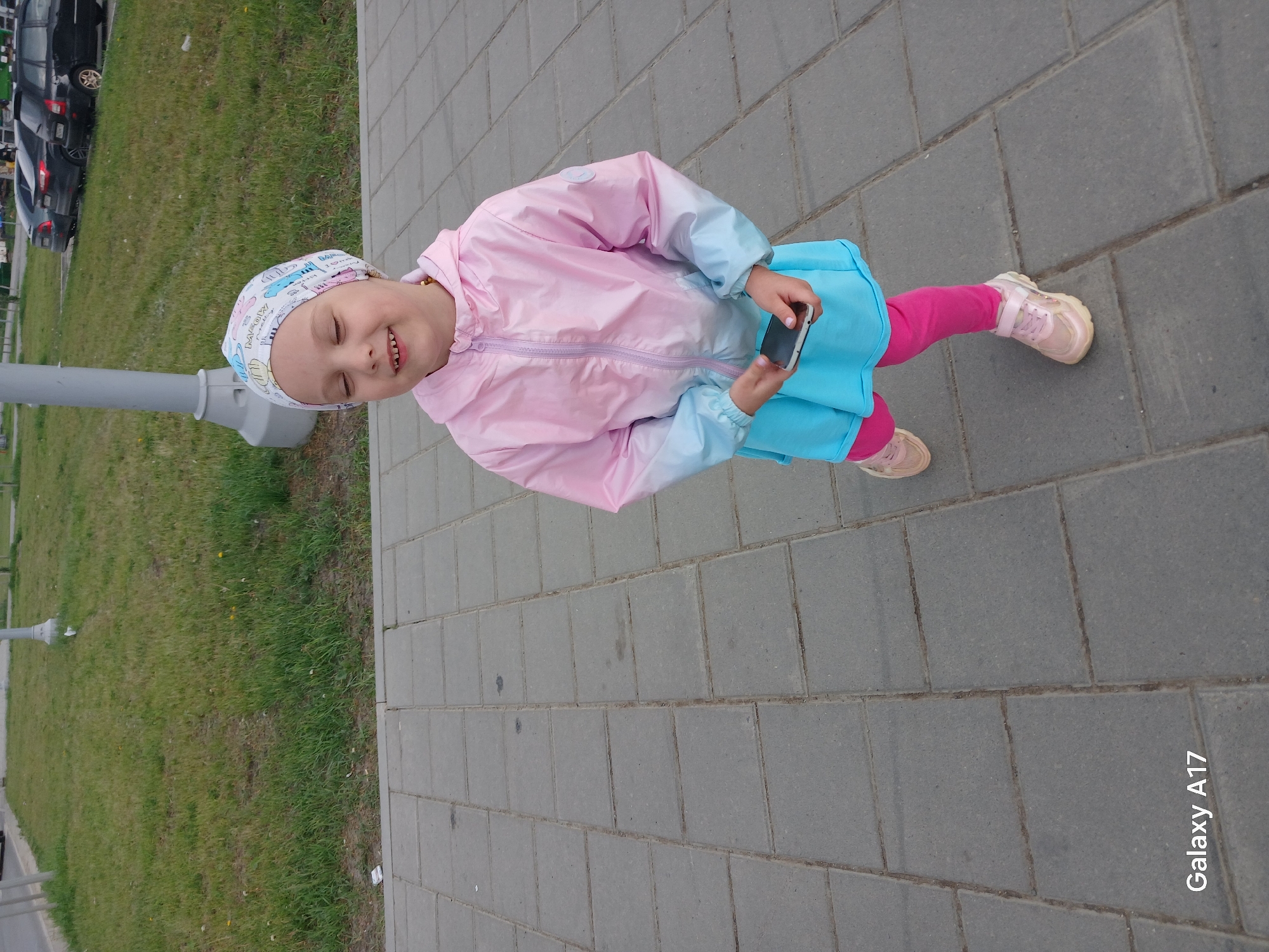 A young child stands on a paved walkway in what appears to be a city park or urban area. The child, with closed eyes and a slight smile, wears a white patterned head covering, a pink and light blue jacket, a blue skirt, and pink leggings. They are holding a smartphone in their hands. The walkway is made of gray pavers, and to the left is a grassy area with a light pole. Cars are visible in the background. The bottom right corner of the image displays the text "Galaxy A17." The scene seems to be in Yekaterinburg, Russia.