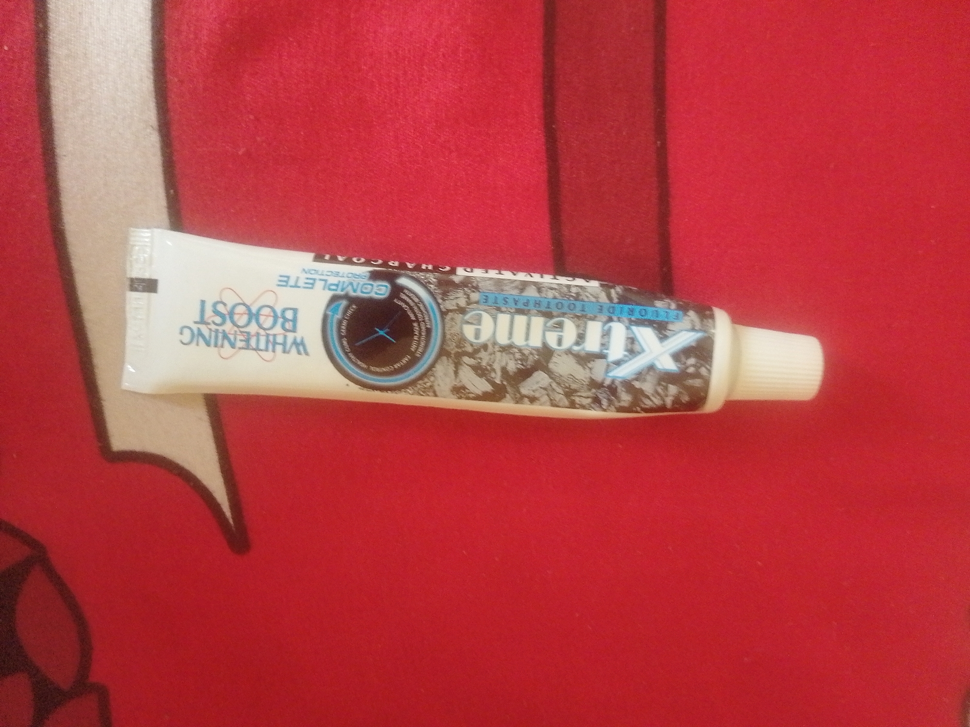A tube of "Xtreme Whitening Boost" toothpaste with activated charcoal and fluoride is positioned horizontally on a red textured surface, possibly a cloth or blanket. The toothpaste tube is white with blue and silver text and graphics. The text "WHITENING BOOST" is prominently displayed in large, bold letters, along with "COMPLETE PROTECTION" and "ACTIVATED CHARCOAL." A circular graphic with a blue border and a stylized clock face or star inside is visible on the tube. The cap of the tube is white. The red surface has a subtle weave pattern. The image is a close-up shot, focusing entirely on the toothpaste tube and the immediate surrounding surface. There is no indication of any specific location within Danbazau, Nigeria, or any other urban environment.