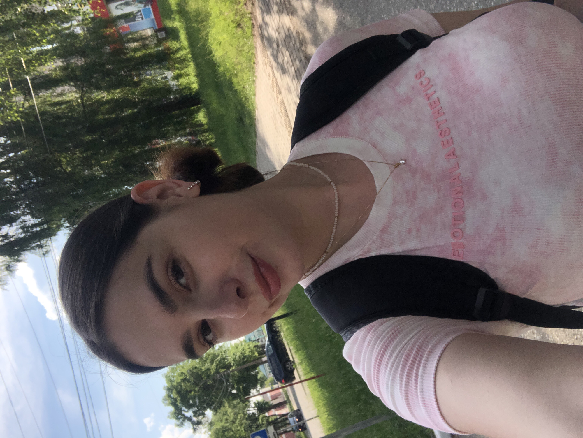 A young woman with dark hair pulled back and a small earring is taking a selfie outdoors. She is wearing a pink tie-dye t-shirt with the words "EMOTIONAL AESTHETICS" printed on it and a black backpack. In the background, there is a tree-lined street with a road, grass, and buildings. The sky is blue with some clouds. A black car and a blue car are visible on the road. The setting appears to be a residential or park area in the city of Chaikovsky, Russia.