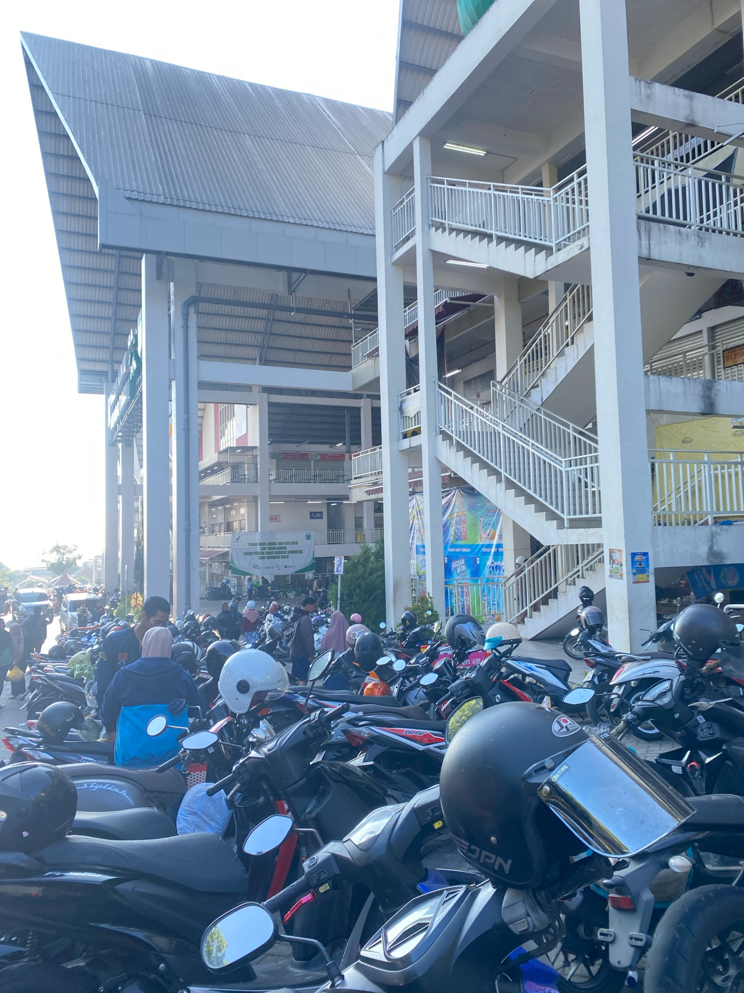 The image depicts a busy outdoor market with a large number of parked motorcycles in the foreground. The market appears to be multi-story with concrete pillars and a large, angled roof structure overhead. Several people are visible, some walking amongst the motorcycles and others standing in groups. One large banner is hanging from a building, displaying text in Indonesian. The sky is bright and appears to be daytime. The overall scene suggests a commercial area likely used for daily shopping.