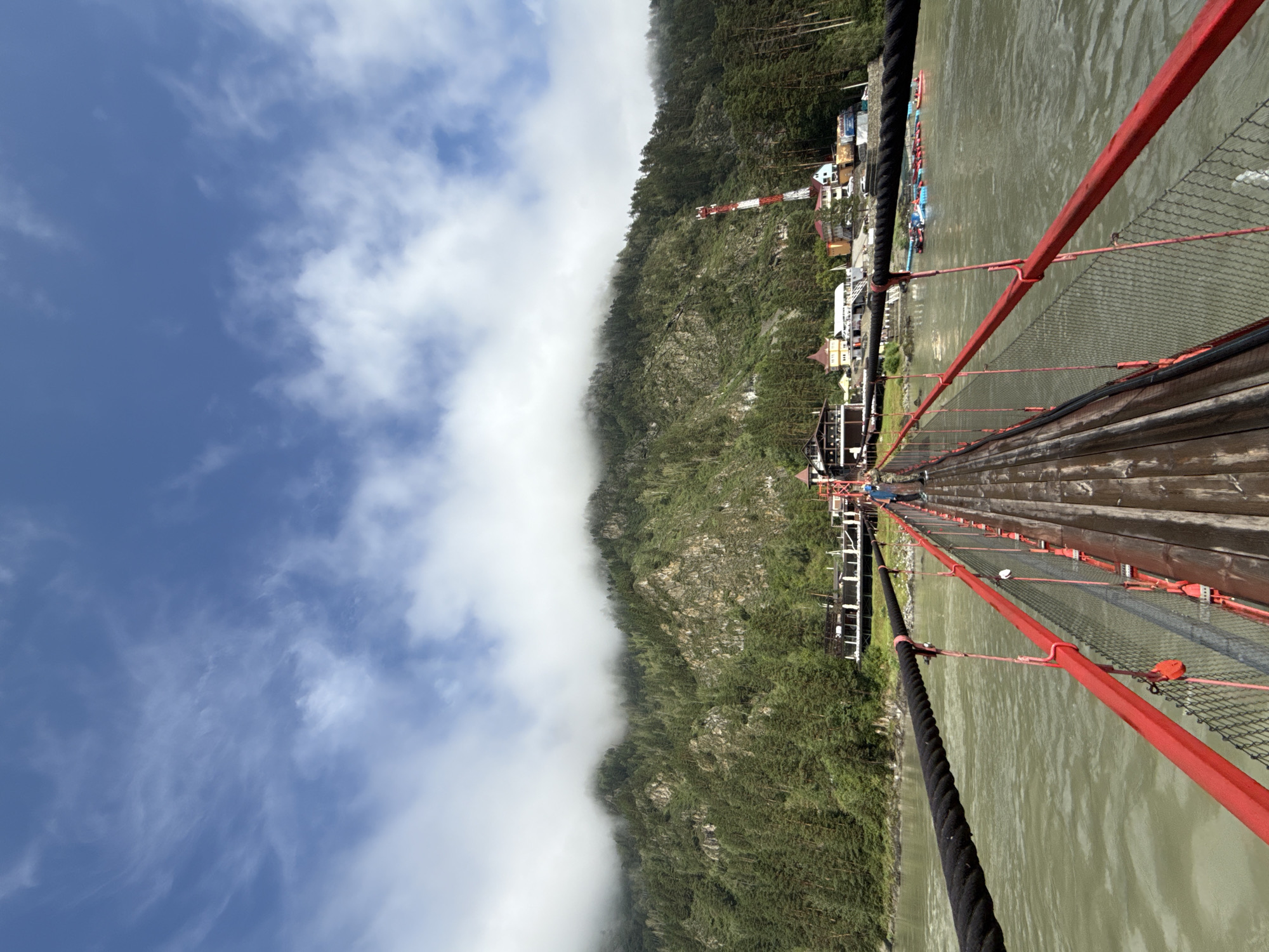 In Barangol, Russia, this image captures a perspective from a wooden suspension bridge. The bridge, with its weathered planks and bright red railings, spans a wide, murky green river. Thick black ropes serve as the main cables supporting the structure. A chain-link fence with red metal poles lines the sides of the bridge for safety. In the distance, the opposite bank is dominated by a steep, densely forested mountain that disappears into a thick layer of fog and clouds clinging to its peak. At the base of the mountain, a cluster of buildings forms a small settlement, including what appears to be a resort or tourist facility with white structures and a prominent red and white communications tower. Several small boats, possibly rafts, are visible in the water near the settlement. A single individual is seen walking across the bridge in the middle distance. The sky above is a vibrant blue, dotted with wispy white clouds.