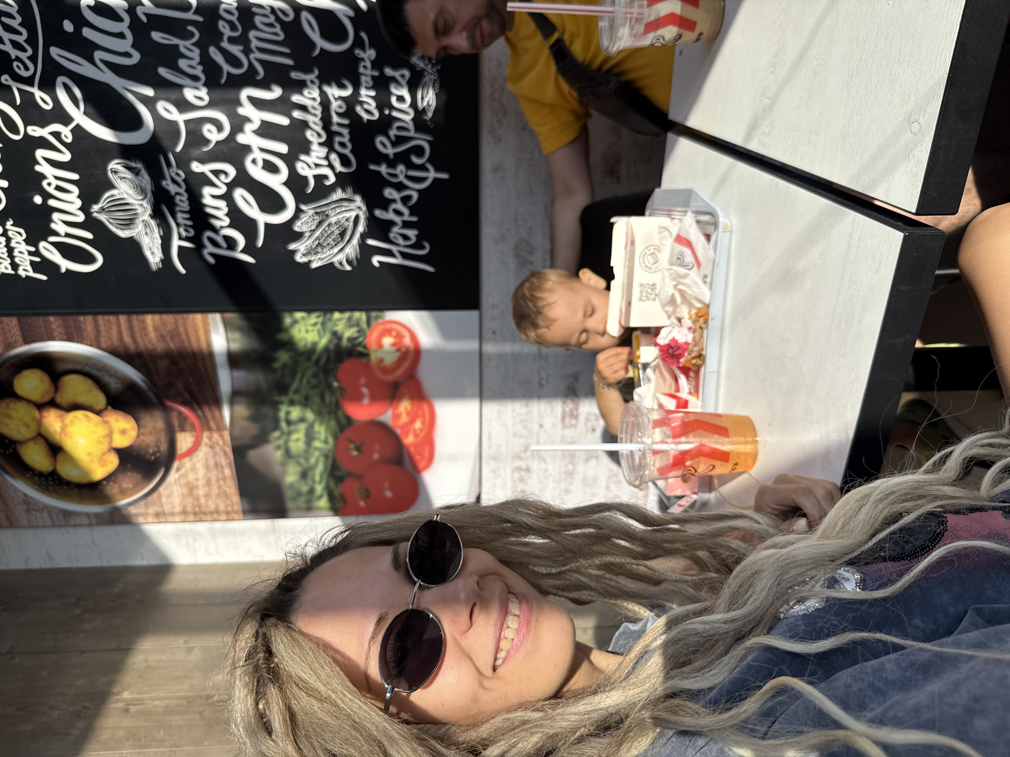 A woman with blonde hair and sunglasses is taking a selfie at an outdoor seating area of a fast-food restaurant. She is wearing a blue denim jacket over a pink shirt. She is smiling and looking at the camera. To her right, a young boy is eating from a fast-food container. A man is seated behind the boy. The table is set with drinks and food wrappers. In the background, there are posters on the wall advertising food items. The scene appears to be in an urban or suburban environment with sunlight casting shadows.