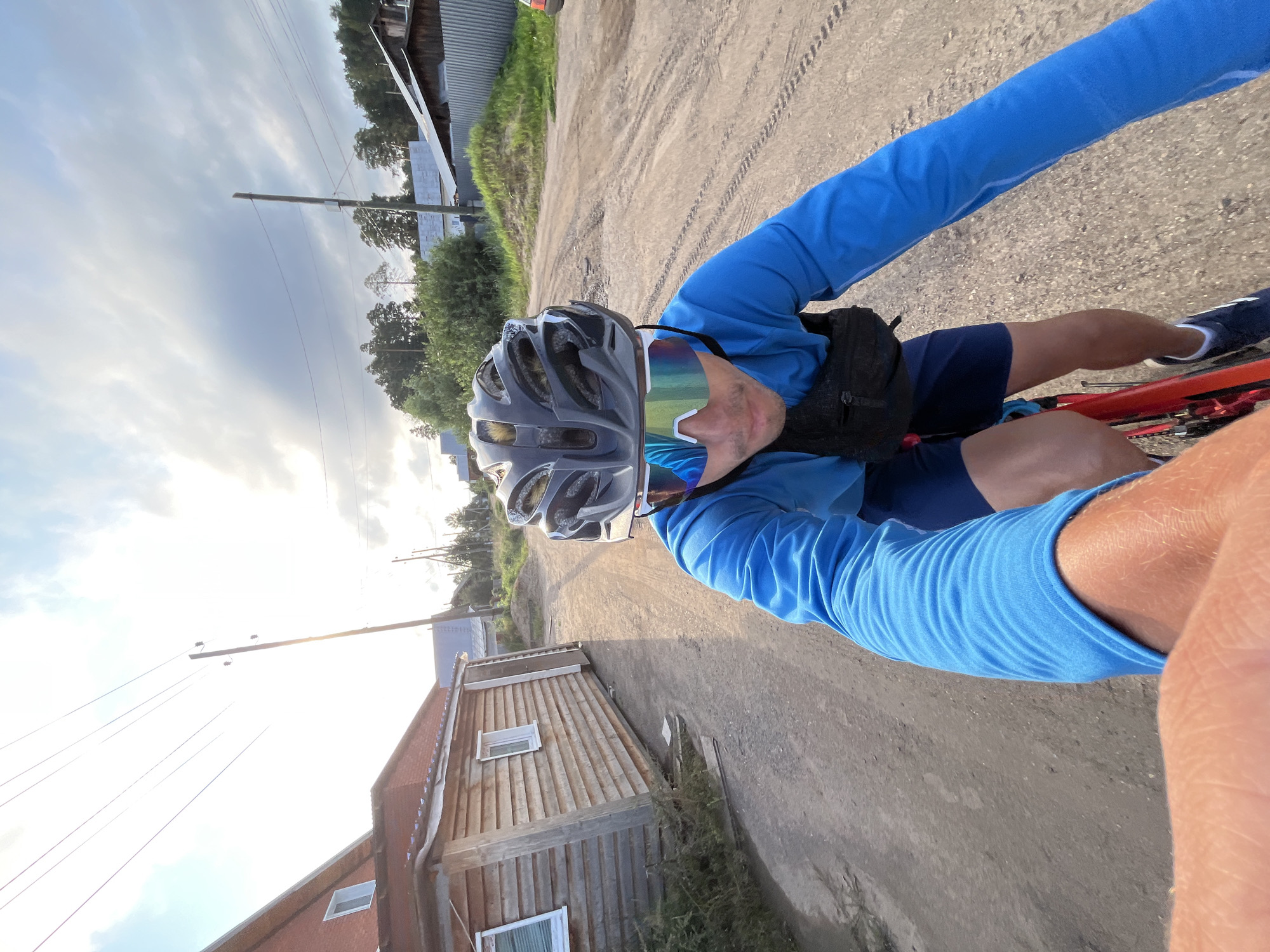 The image shows a first-person perspective of a person cycling on a dirt road. The cyclist is wearing a blue long-sleeved shirt, dark blue shorts, a gray helmet, and iridescent cycling glasses. A black fanny pack is worn around their waist. The road appears dry and dusty, with tire tracks visible.
To the left of the road, there are two wooden buildings. The building closer to the foreground is a single-story structure with a reddish-brown metal roof and light-colored wooden siding. It has two windows visible. Further in the background, another building with a grey corrugated metal roof and white siding is partially visible. Beyond the buildings, there are trees and more distant structures. Overhead, power lines are strung between wooden poles, and the sky is partly cloudy with the sun visible, suggesting it is either morning or late afternoon. The overall lighting indicates a bright day.
The setting appears to be a rural or semi-rural area, consistent with the context of Chaikovsky, Russia. The cyclist's posture and the motion blur suggest they are actively riding. No visible text is present in the image.