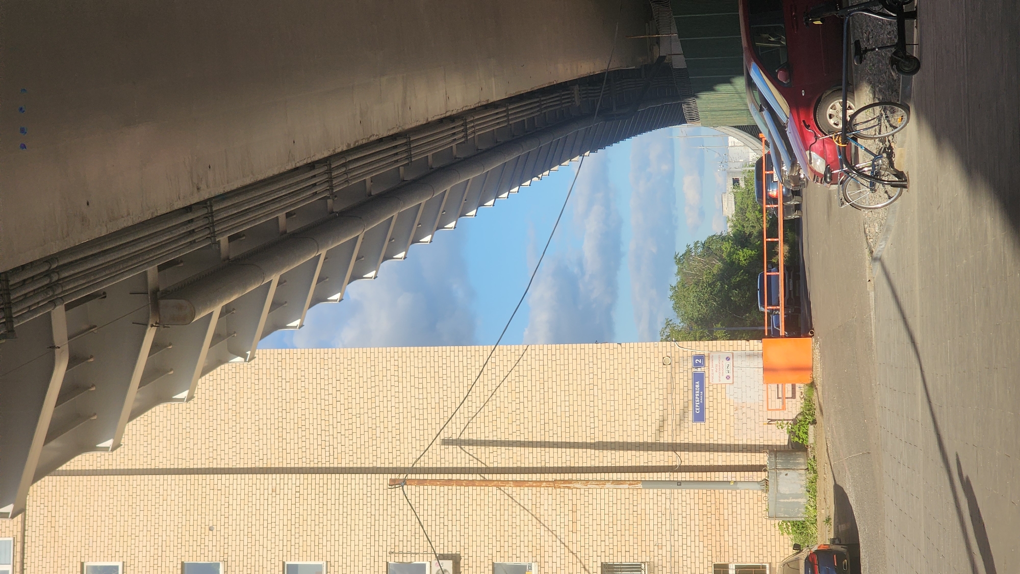 The image shows a street scene in Moscow, Russia. A brick building with windows runs along the lower part of the frame. Above, a concrete overpass structure creates a sense of enclosure. The sky is visible between the building and the overpass, revealing clouds and blue sky. A street sign with Cyrillic text that reads "СЕРЕБРЯКОВА 2" (Serebryakova 2) is affixed to the brick building. On the right side of the image, there is a street with a car parked on it and a bicycle leaning against the car. An orange barrier is visible near the building's entrance. The lighting indicates it is daytime.