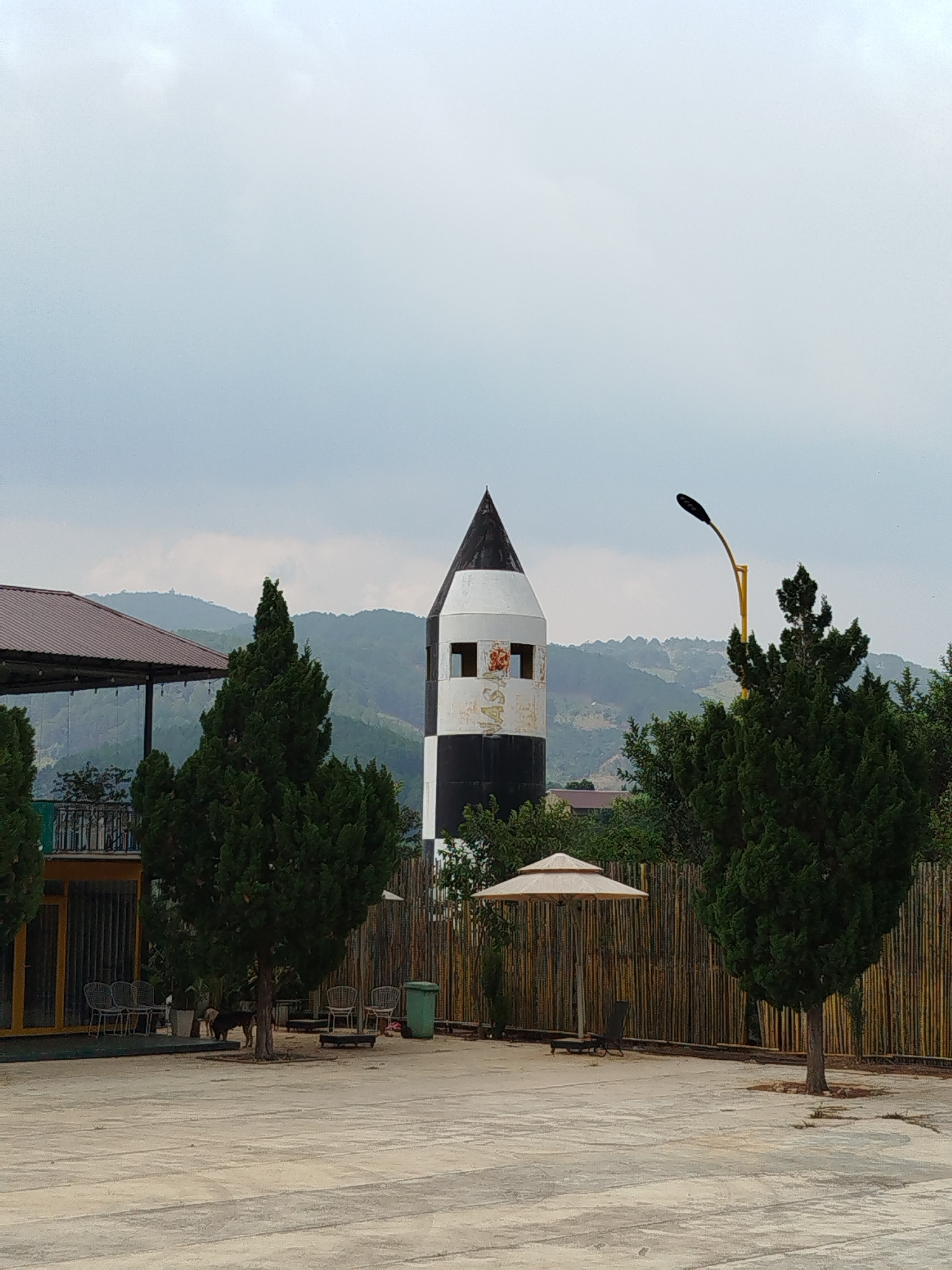 The image captures an outdoor scene at what appears to be a resort or recreational area, set against a backdrop of rolling hills under an overcast sky. In the center, a striking black and white tower with a conical black top dominates the mid-ground. This tower features several rectangular openings or windows; one of these clearly displays white letters on a red background, appearing to spell "WASI" or similar.
To the left, a building with a dark brown, tiled roof and an open-air structure is partially visible, with support pillars and railings. Numerous tall, dark green coniferous trees are scattered throughout the foreground and mid-ground, framing the scene. A bright yellow street lamp with a curved arm and a dark fixture stands to the right of the tower.
The foreground consists of a large, light-colored paved or concrete area with faint patterns, suggesting a courtyard or plaza. In the mid-ground, there's a light-colored outdoor umbrella with an open canopy and a lounge chair beneath it, positioned near a tall bamboo fence. Under one of the trees on the left, several outdoor chairs and tables are arranged, and at least one dog, possibly two, can be seen standing near the base of the tree. A green trash can is also visible near the bamboo fence. The distant background features multiple layers of green mountains or hills, partially obscured by the hazy, light grey sky.
The scene depicts a calm daytime setting, likely under cloudy conditions given the diffused lighting and lack of sharp shadows. No human activity is overtly visible, suggesting a tranquil moment at the location. The overall impression is one of an inviting outdoor space.