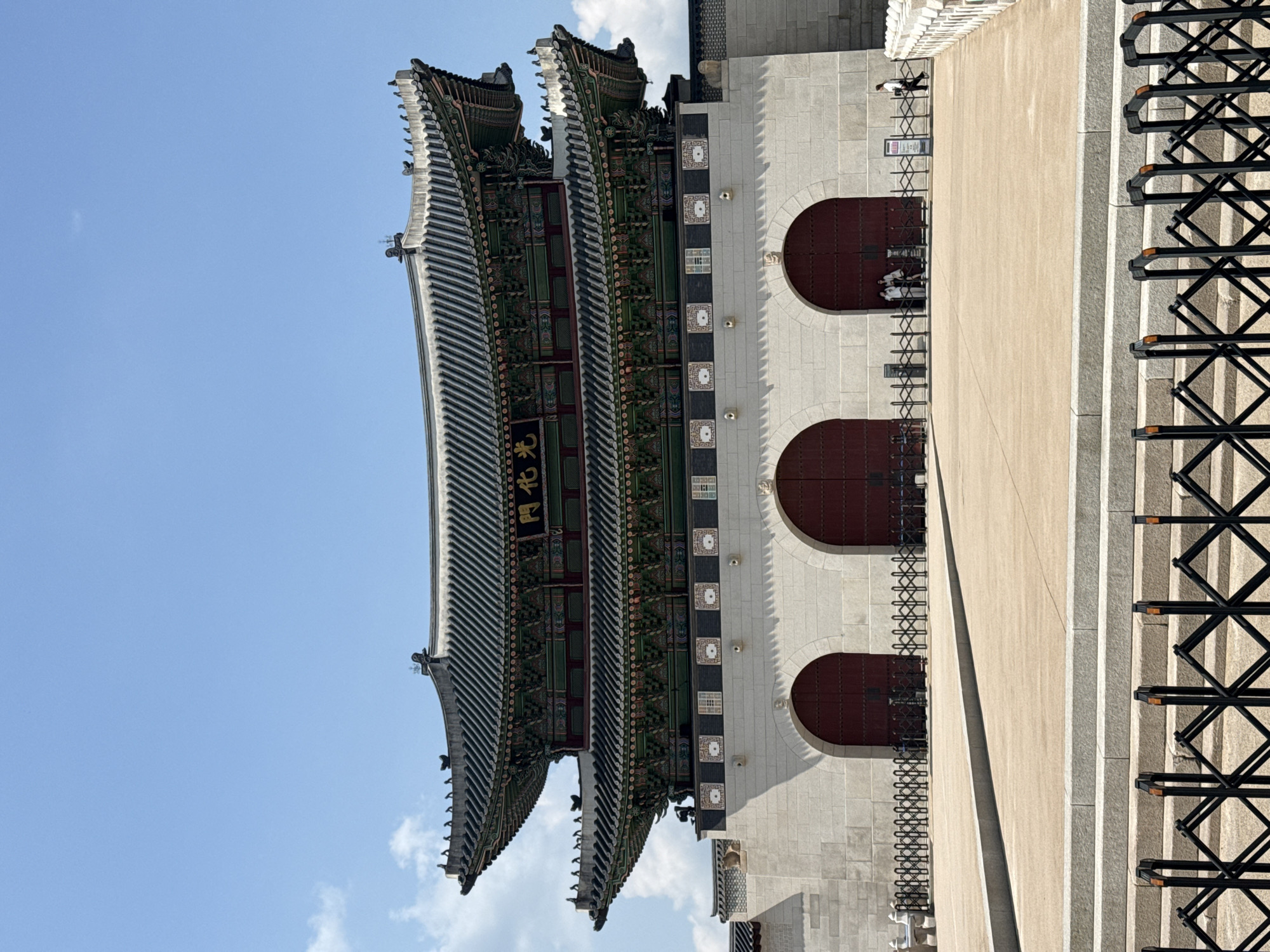 The image shows a building in Seoul, South Korea, specifically the Gwanghwamun Gate at Gyeongbokgung Palace. The gate is a large, ornate structure with a multi-tiered roof and decorative elements. The wall of the gate is made of white stone, and there are three arched doorways in the facade. Black metal crowd barriers are visible in the foreground on the right. A few people are near the gate, but their actions and interactions are not visible. The sky is blue with a few clouds.