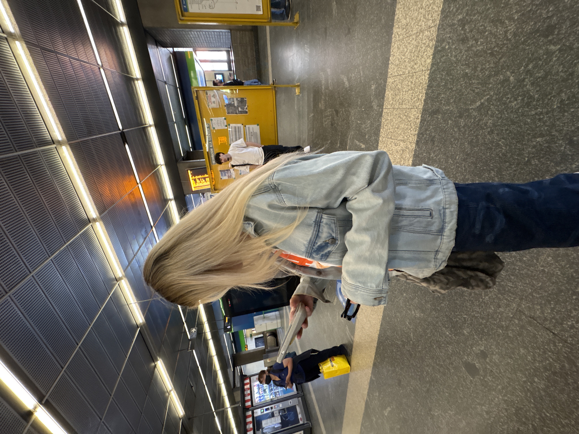 The image shows a passenger in the Milan Metro, Italy. The passenger has blonde hair and is wearing a denim jacket and blue jeans. They are looking at a phone. A person is sitting near a yellow bag in the background. Two people wearing face masks stand in front of ticket vending machines. Another person is lying down near a yellow public information board. The station has gray floors, gray acoustic panels on the ceiling, and bright fluorescent lighting.