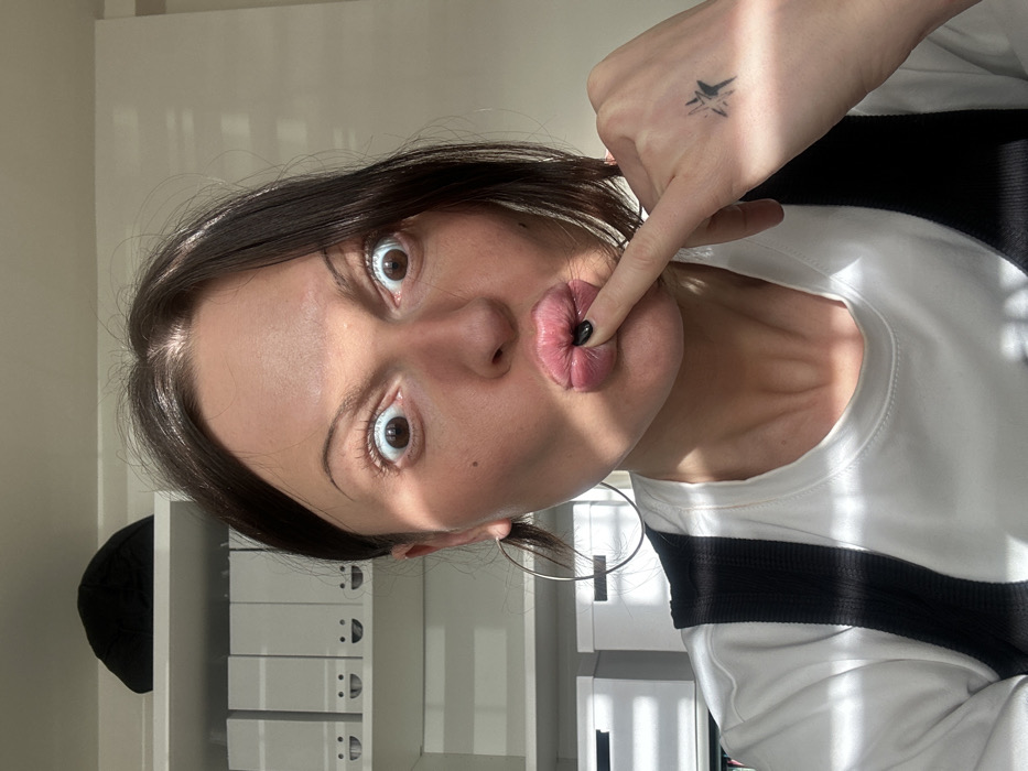 An adult female is depicted in a brightly lit indoor close-up, likely a selfie. She has dark brown hair and is wearing a white t-shirt with a dark strap over it, and a large silver-colored hoop earring on her left ear. Her face exhibits an exaggerated, playful expression with wide-open eyes showing significant white sclera, raised eyebrows, and tightly puckered lips. Her left index finger, with a black-painted nail, is placed against the center of her puckered lips. A small, black, stylized star or cross tattoo is visible on the back of her left hand.
The background, largely out of focus, reveals a white shelving unit. On the top shelf, a dark soft object, possibly a cap, is visible. On the shelf below it, several identical white document boxes are arranged, each featuring a simple black smiley face drawn or printed on its front. More white storage boxes are partially visible on a lower shelf to the left. Strong natural light originates from the viewer's right, casting distinct shadows across the subject's face and the background. The scene is located indoors in Tunbridge Wells, United Kingdom.