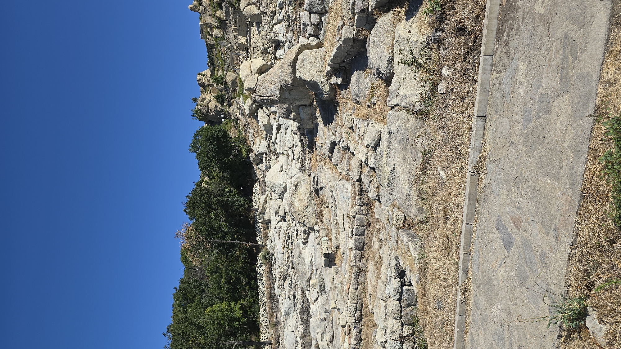 The image depicts a view of the ancient Thracian city of Perperikon, located near the modern city of Gorna Krepost, Bulgaria. The scene shows the archaeological site under a clear blue sky. In the foreground, there is a paved pathway made of irregular stone slabs, bordered by dry, grassy vegetation. The pathway leads upwards towards the ruins. The main focus of the image is the extensive ruins themselves, which consist of numerous large, scattered stone blocks, remnants of ancient structures, and visible foundations constructed from smaller stones. These ruins are situated on a rocky hillside. In the background, a dense cluster of green trees can be seen on the upper slope of the hill, and further up, more rocky outcrops and stone structures indicative of the ancient city are visible. The overall impression is one of a historical site exposed to the elements, with the natural landscape interacting with the man-made ruins.