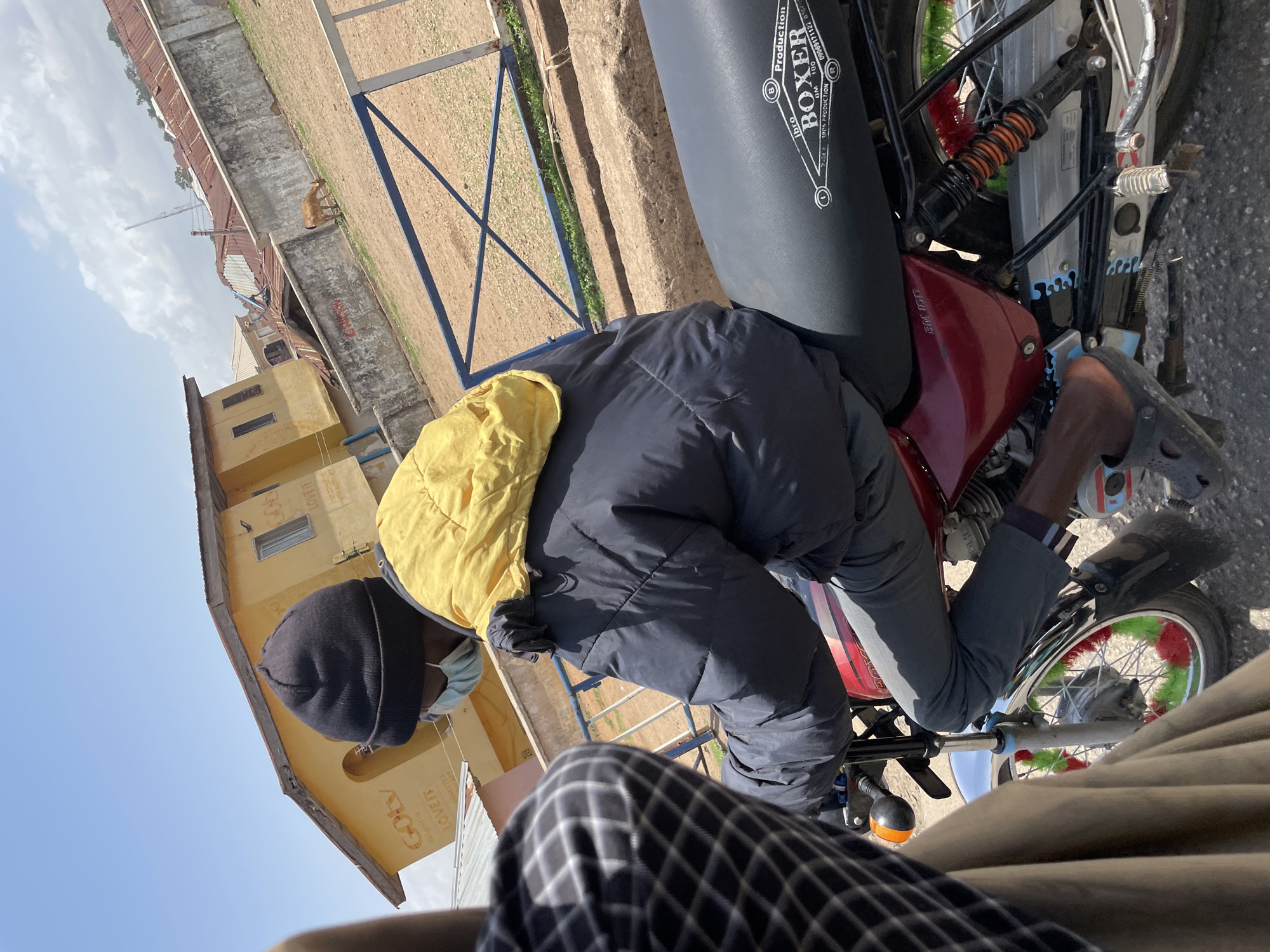 A person, wearing a black beanie, a light blue face mask, a dark puffy jacket with a yellow hood draped over it, and grey pants, is riding a red and black motorcycle. The motorcycle has "BOXER" and "Production" written on the seat. The person is wearing dark colored clogs on their feet. The motorcycle's front wheel has red and green decorations on the spokes. In the background, there is a yellow building with text that appears to read "Gbay" and "LOVE IT". Further in the background, there are other buildings with corrugated metal roofs, a concrete wall, a dirt ground, and a blue fence. A brown goat is visible grazing near the concrete wall. The sky is clear and blue with some clouds. This scene is located in Jos, Nigeria.