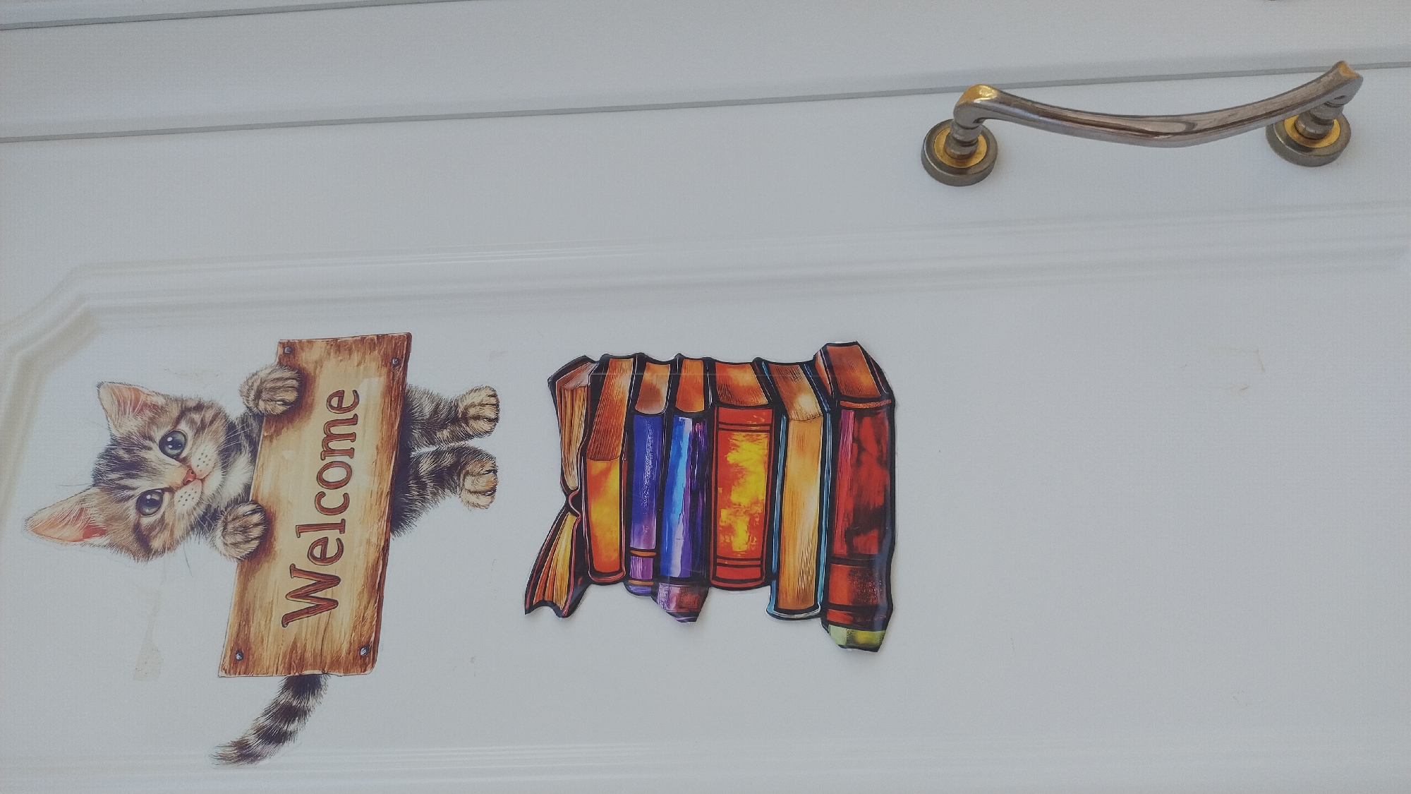 A decorative sticker of a kitten holding a "Welcome" sign and a stack of colorful books is affixed to a white door. The kitten is depicted with tabby markings, large eyes, and is positioned above the stack of books. The books are arranged in a slightly offset, vertical pile, showcasing spines in shades of red, orange, yellow, blue, and purple. The door has a visible handle on the right side, which is metallic and curved. The scene appears to be on the exterior of a building. The provided contextual information states this image is from Νέα Μηχανιώνα, Greece.
