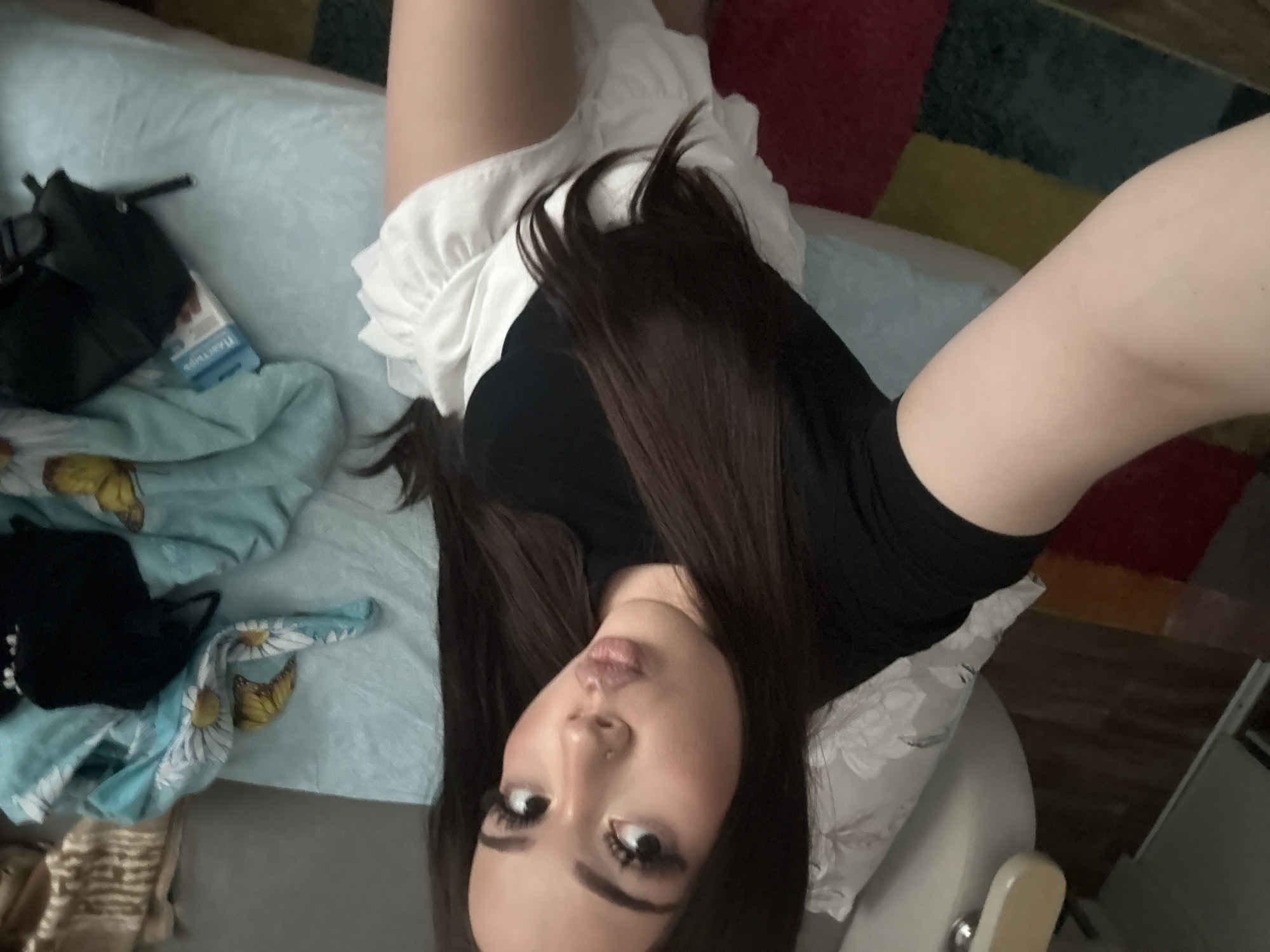 The image depicts a young woman with long, dark hair, wearing a black top and a white ruffled skirt. She is posing for a selfie, puckering her lips. The background suggests an indoor setting, possibly a bedroom. A light blue blanket with a floral and butterfly pattern is visible, along with some clothing items. A small blue box, possibly containing medication or personal care items, and a black bag are also present. The floor is covered with a colorful rug. The image does not provide any information about the location within Naberezhnye Chelny, Russia, or any specific actions or interactions.