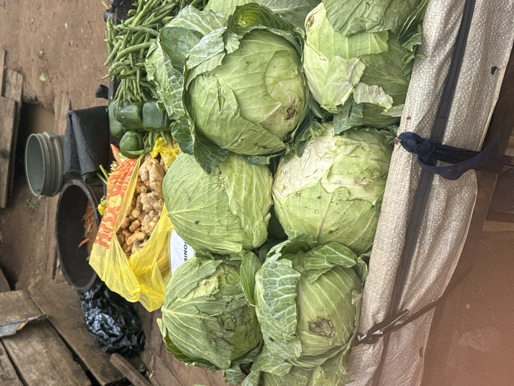 This is a photograph taken at a market stall in Ihiagwa, Nigeria. The stall is laden with fresh produce, predominantly large, green cabbages. In the foreground, several cabbages are piled on top of a beige, textured tarp that appears to be part of a stall structure. A black drawstring is visible, tied to the tarp.
Behind the cabbages, a bright yellow plastic bag is filled with what appears to be ginger or turmeric roots. To the left of the yellow bag, a black plastic bag is visible, and further left, a black basin contains some red chili peppers and other items. In the background, a pile of fresh green beans is arranged next to some green bell peppers. The ground appears to be dirt. The overall impression is one of a vibrant, bustling local market scene.