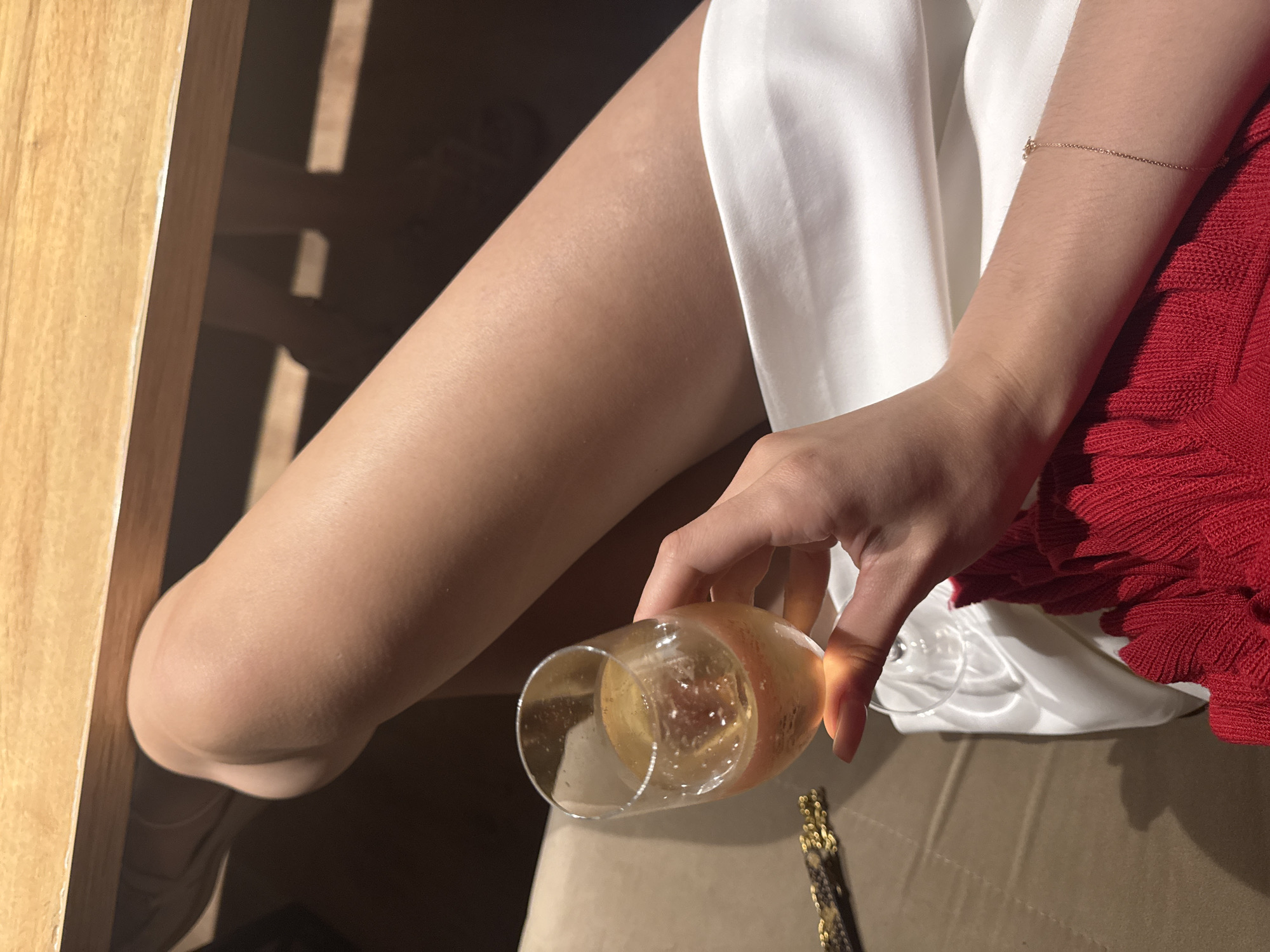 The image depicts a close-up view of a woman's legs and arm, holding a glass of champagne. She is seated at a table, with her legs crossed and draped over the edge. The background shows a wooden table and what appears to be a neutral-colored sofa or chair. The woman is wearing a red garment and a white fabric. A thin gold bracelet is visible on her wrist. The setting is not identifiable as being in the city of Chaikovsky, Russia.