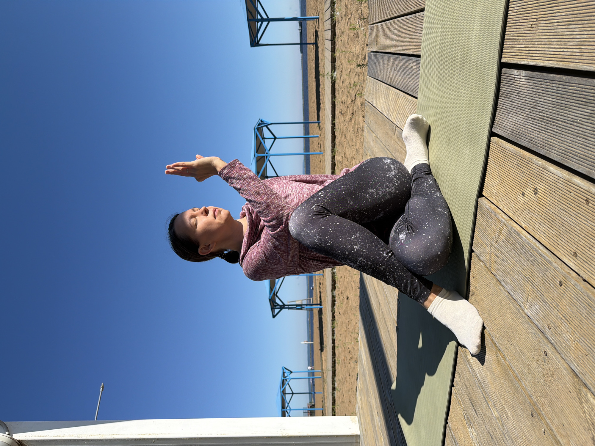A woman is performing a yoga pose on a mat outdoors on a wooden deck. She is wearing a pink sweater and black patterned leggings. Her eyes are closed and her right hand is raised with two fingers extended. The background shows a sandy beach with blue structures and a body of water under a clear blue sky. This scene appears to be on the embankment of the Kama River in Chaikovsky, Russia, given the specific blue canopy structures often found in Russian parks and recreation areas, and the presence of the river.