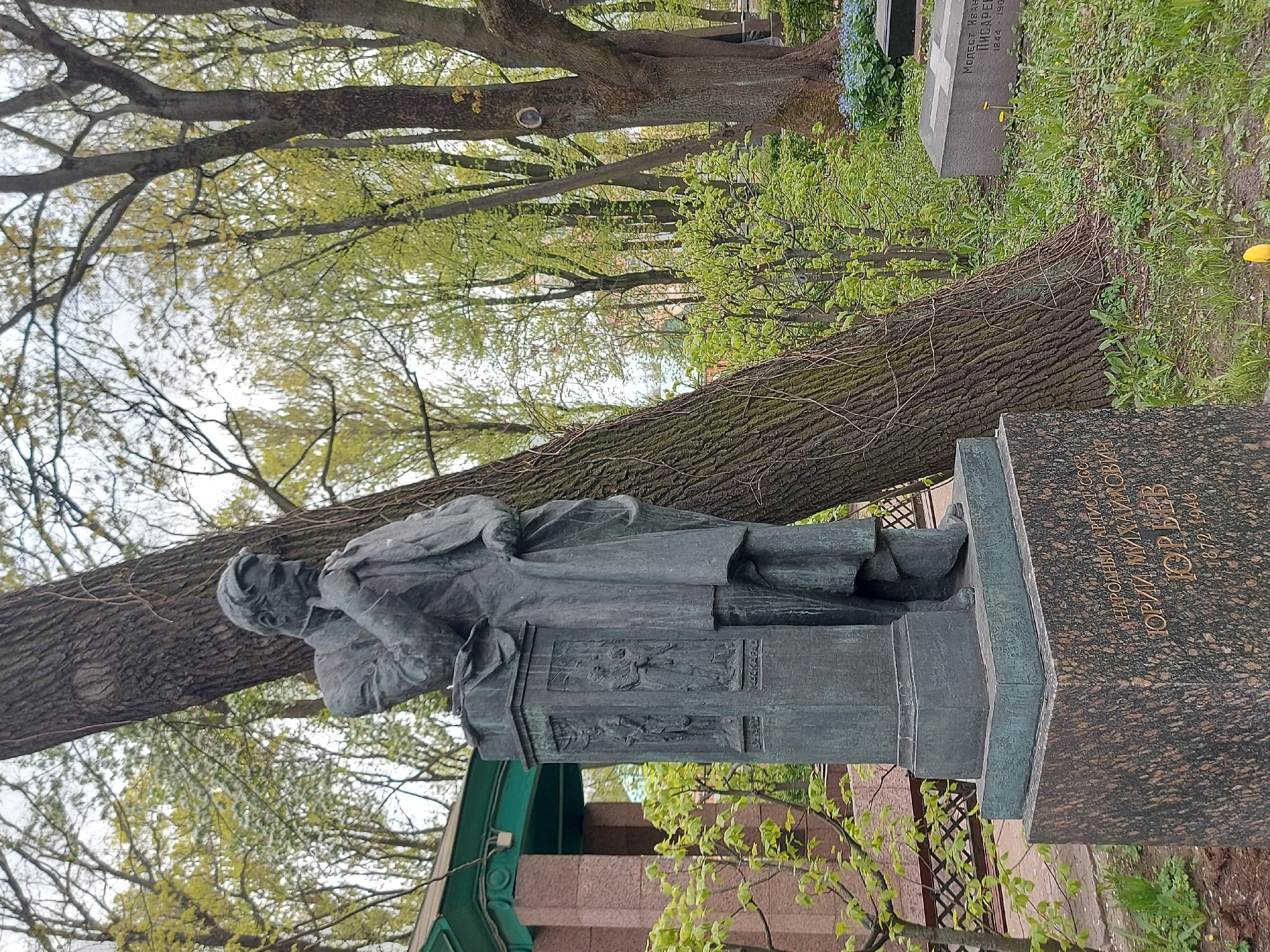The image shows a statue of a man in a long coat standing on a pedestal in a cemetery. The pedestal has two relief panels. The statue is in front of a large tree. To the right of the statue is a gravestone with Cyrillic text. The grave belongs to Russian artist Yuri Mikhailovich Yuryev. The statue is at the Literatorskie Mostki, a section of the Volkovo Cemetery in Saint Petersburg, Russia. There are trees, grass, and other gravestones in the background.