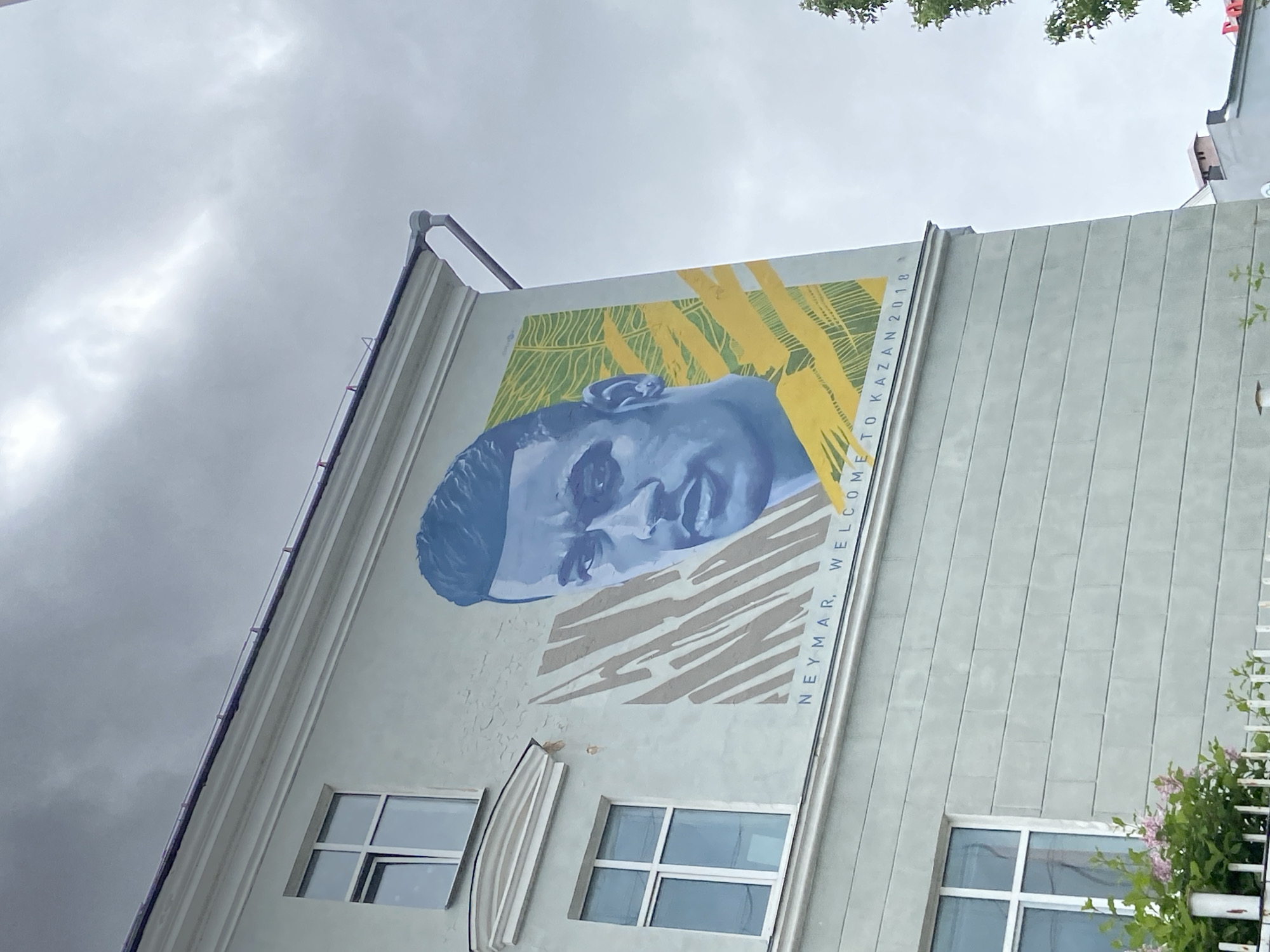An eye-level, tilted shot captures a large mural of the Brazilian football player Neymar on the side of a light-colored building in Kazan, Russia. The mural, rendered in shades of blue for the face, features a background of yellow and green stylized leaf or plant patterns. To the right of Neymar's face, vertical text reads "NEYMAR, WELCOME TO KAZAN 2018," indicating its creation for the 2018 FIFA World Cup hosted in Russia.
The building itself is light grey or off-white, with a textured facade. Several rectangular windows with white frames are visible in the lower part of the wall. A dark rain gutter or pipe runs along the upper left edge of the building. The sky above is heavily overcast and cloudy, suggesting a gray, possibly damp, day. On the far right, some green foliage with small pink flowers adds a touch of natural color. The overall scene depicts urban art commemorating a significant sporting event, set against a muted sky and functional architecture.