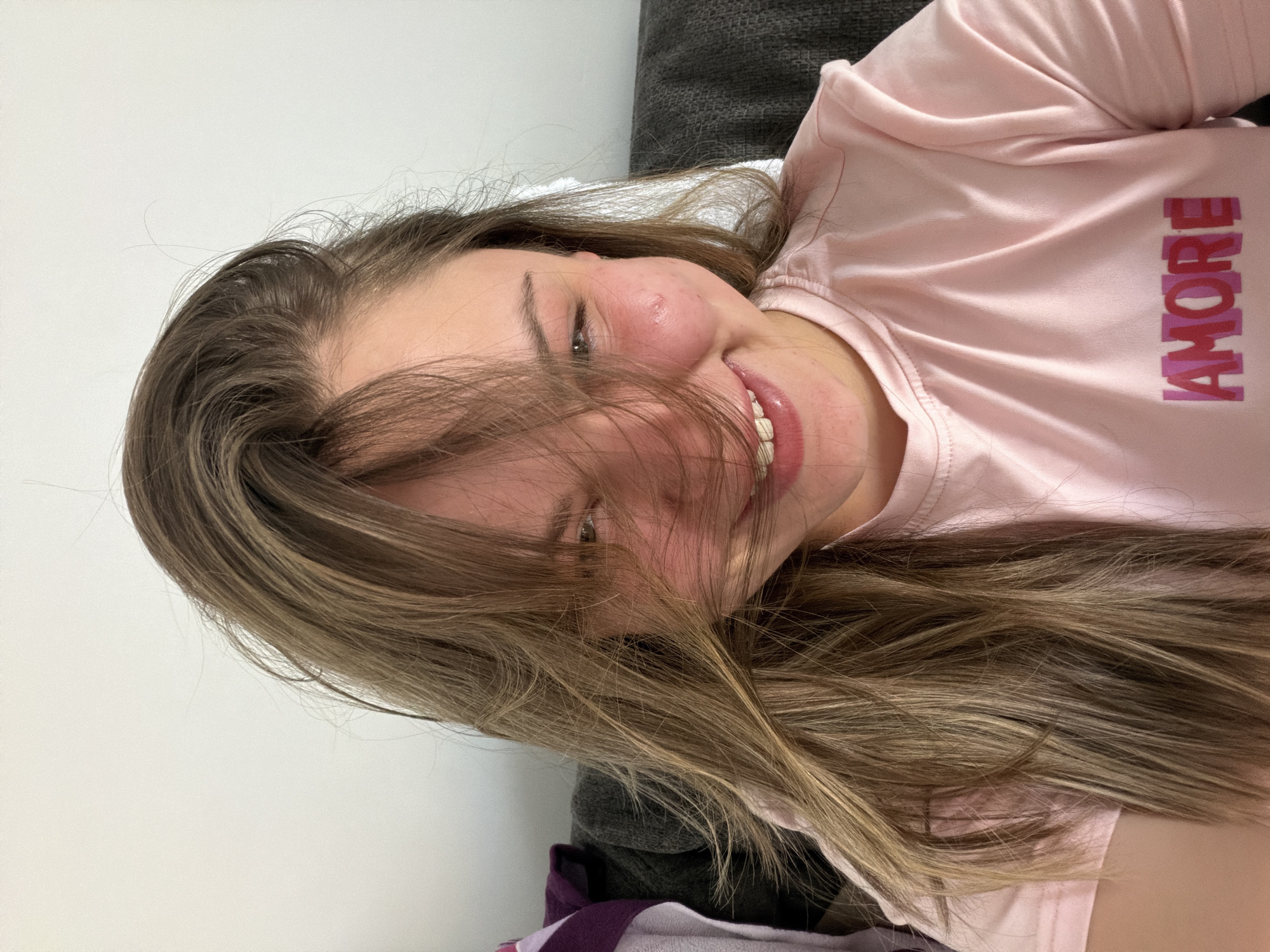 A young woman is smiling while lying on a gray couch. She is wearing a pink shirt with the word "AMORE" written on it. Her brown and blonde hair is partially covering her face. The background includes a white wall. There is no information in the image to place it in the city of Arroyo de la Miel-Benalmádena Costa, Spain.