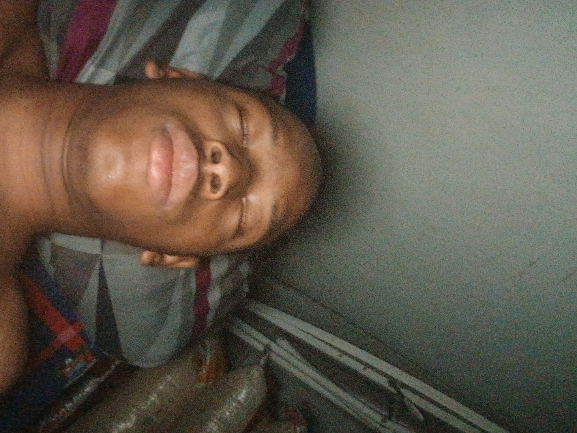 A person with dark skin lies on their back with eyes closed. They are on a bed with a patterned blanket. A wall with a few horizontal lines runs alongside the bed. The location is indeterminable.