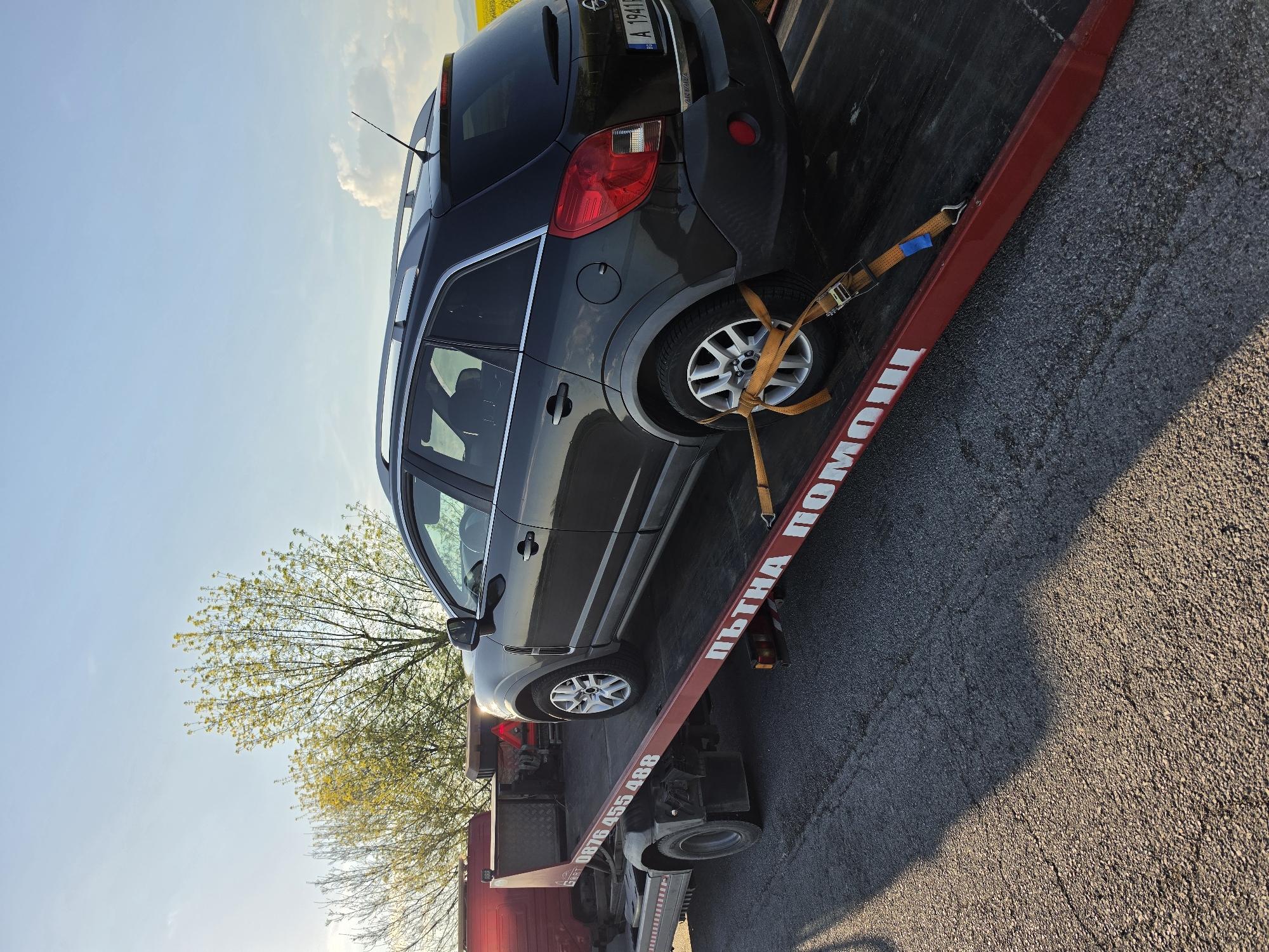 A dark grey Opel Antara is secured with yellow straps onto a red flatbed tow truck, with the Bulgarian phrase "ПЪТНА ПОМОЩ" in white letters on the side of the flatbed. The license plate of the car is visible. The tow truck has the phone number "0876 455 488" and the word "GART" on the bottom. The scene appears to be on an asphalt road, with a field or trees in the background under a blue sky with light clouds. Based on the provided information, this likely took place in or near Polsko Padarevo, Bulgaria.