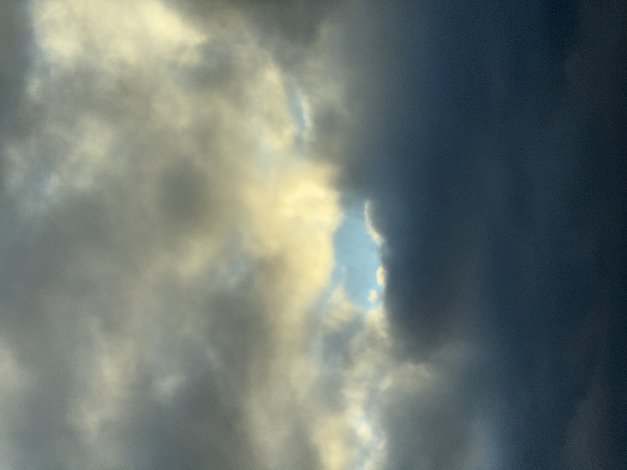 A photograph captures a cloudy sky with a patch of blue visible through a gap in the clouds. The clouds are a mix of dark gray and lighter, yellowish-white hues, suggesting either a stormy or overcast day with some sunlight breaking through. The image is devoid of any identifiable landmarks or structures that would indicate a specific location within Tallinn, Estonia. There are no people, subjects, or objects visible, and no actions or interactions are depicted. The focus is solely on the atmospheric conditions of the sky.