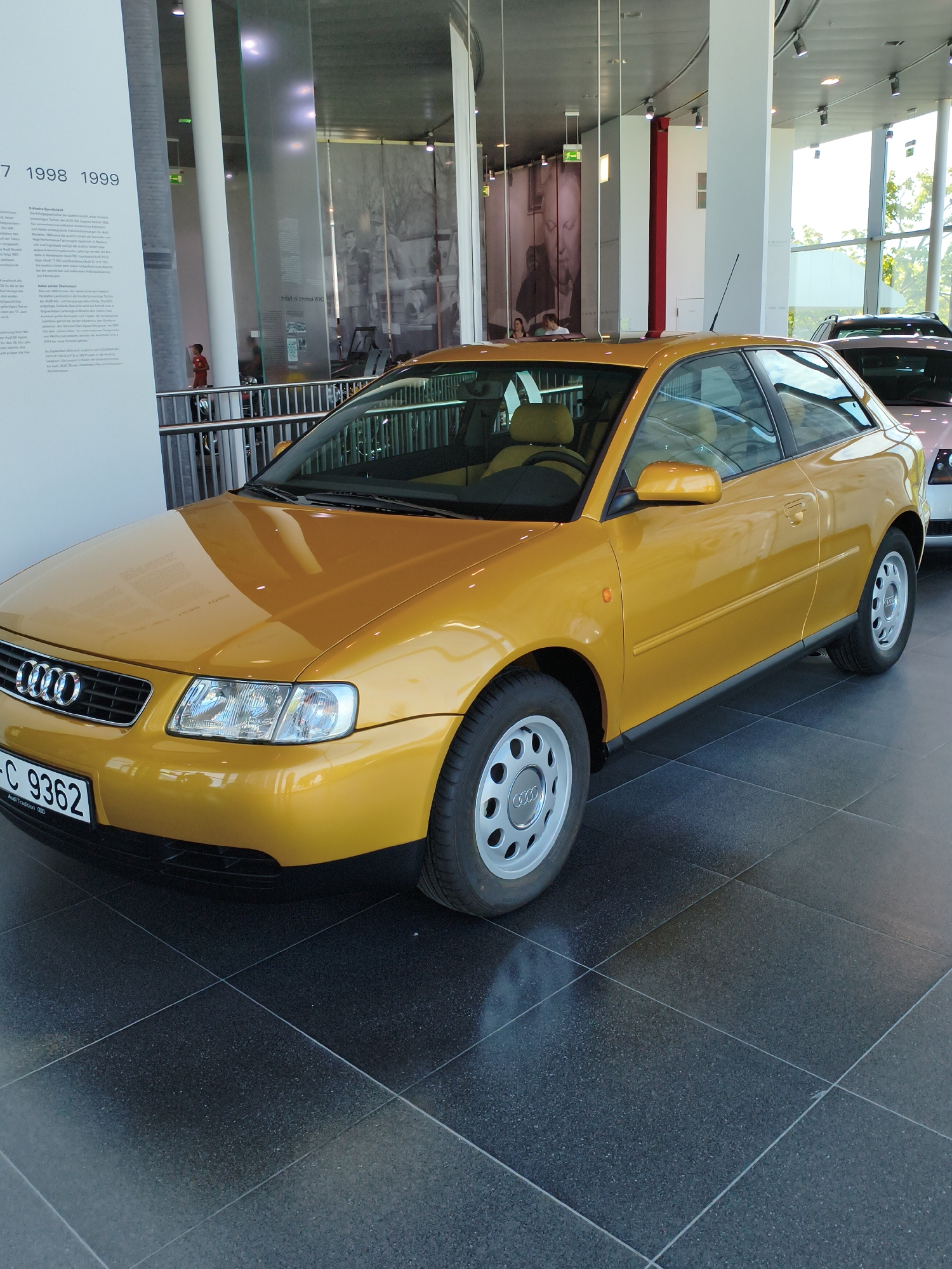 In the Audi Forum Ingolstadt museum, a bright yellow Audi A3 two-door hatchback is displayed on a dark tiled floor. The car, manufactured in 1999, is a prominent exhibit. The background reveals a modern, open-plan exhibition space with large windows offering a view of trees outside. Vertical banners with historical information and images related to Audi's history are visible along a wall, suggesting a curated display. A silver fence separates the exhibit area from a walkway where a person in a red shirt is briefly seen walking. The lighting is bright, illuminating the car and the surrounding environment, highlighting the sleek lines of the vintage vehicle.