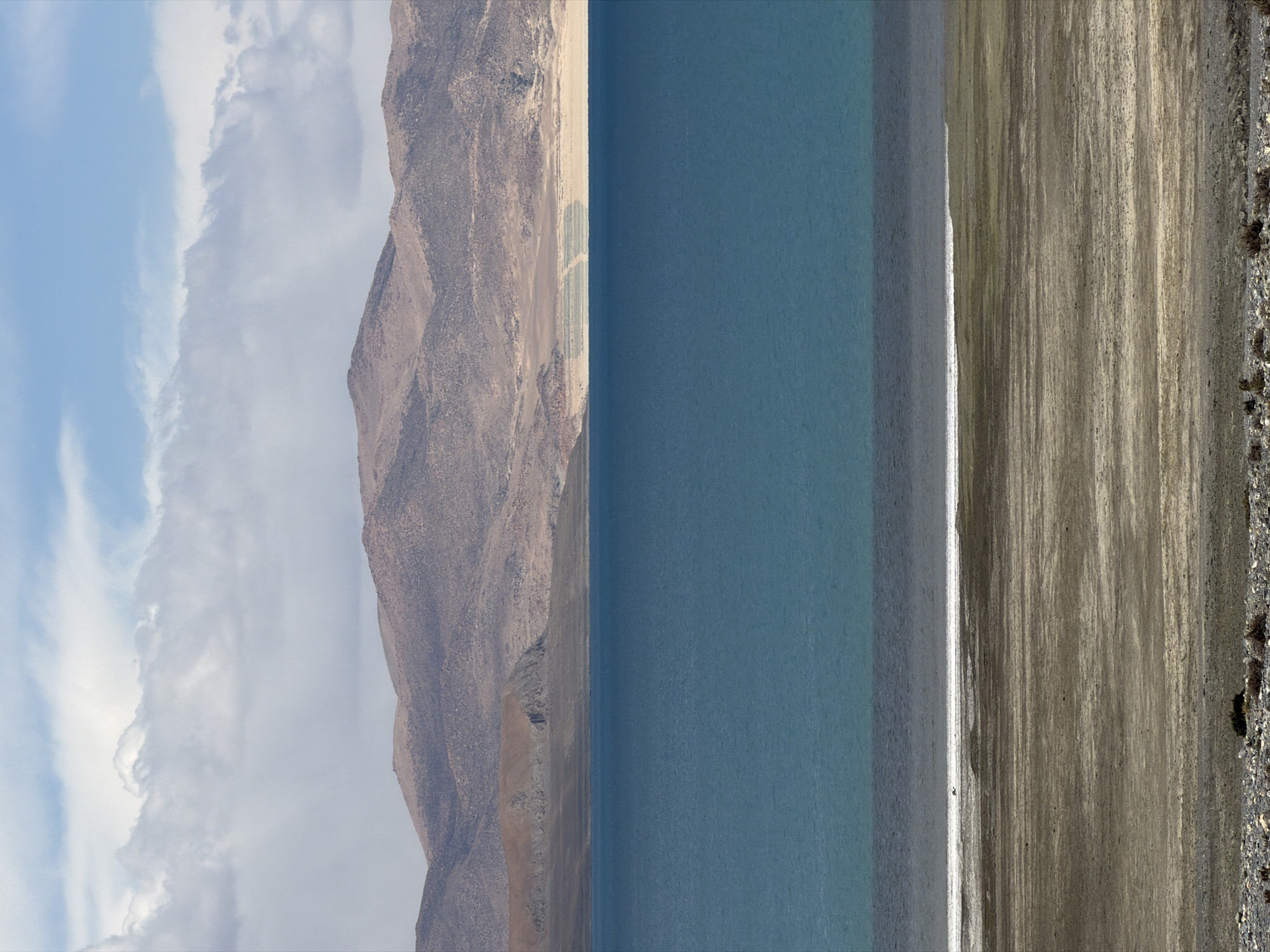 The image shows a landscape in Gyirong County, China. The scene is vertically oriented, with the sky and clouds on the left, transitioning into a tan mountain range. Adjacent to the mountains is a body of turquoise water. To the right of the water is a shoreline with sandy, earthy tones, and a border of small rocks and sparse vegetation along the right edge of the image.