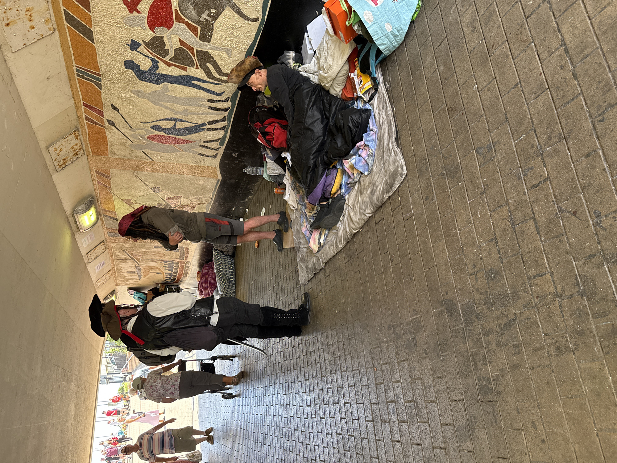 In St Leonards, United Kingdom, a pedestrian underpass has a section designated for rough sleeping. The underpass has a tiled floor and a mural depicting figures in procession on the right wall. In the foreground, a person is sleeping in a black sleeping bag on a mat surrounded by blankets and belongings, including a red backpack and a plastic bottle of water. To the left, two individuals dressed as pirates are standing, facing towards the sleeping person. One pirate, wearing a black leather vest over a white shirt and tall black boots, has a sword at his hip. The other pirate, wearing a green jacket and a red headscarf, stands with his arms crossed. Beyond them, the underpass opens to daylight where other pedestrians are visible, including a man in a striped t-shirt and shorts, and a woman with a walker.
