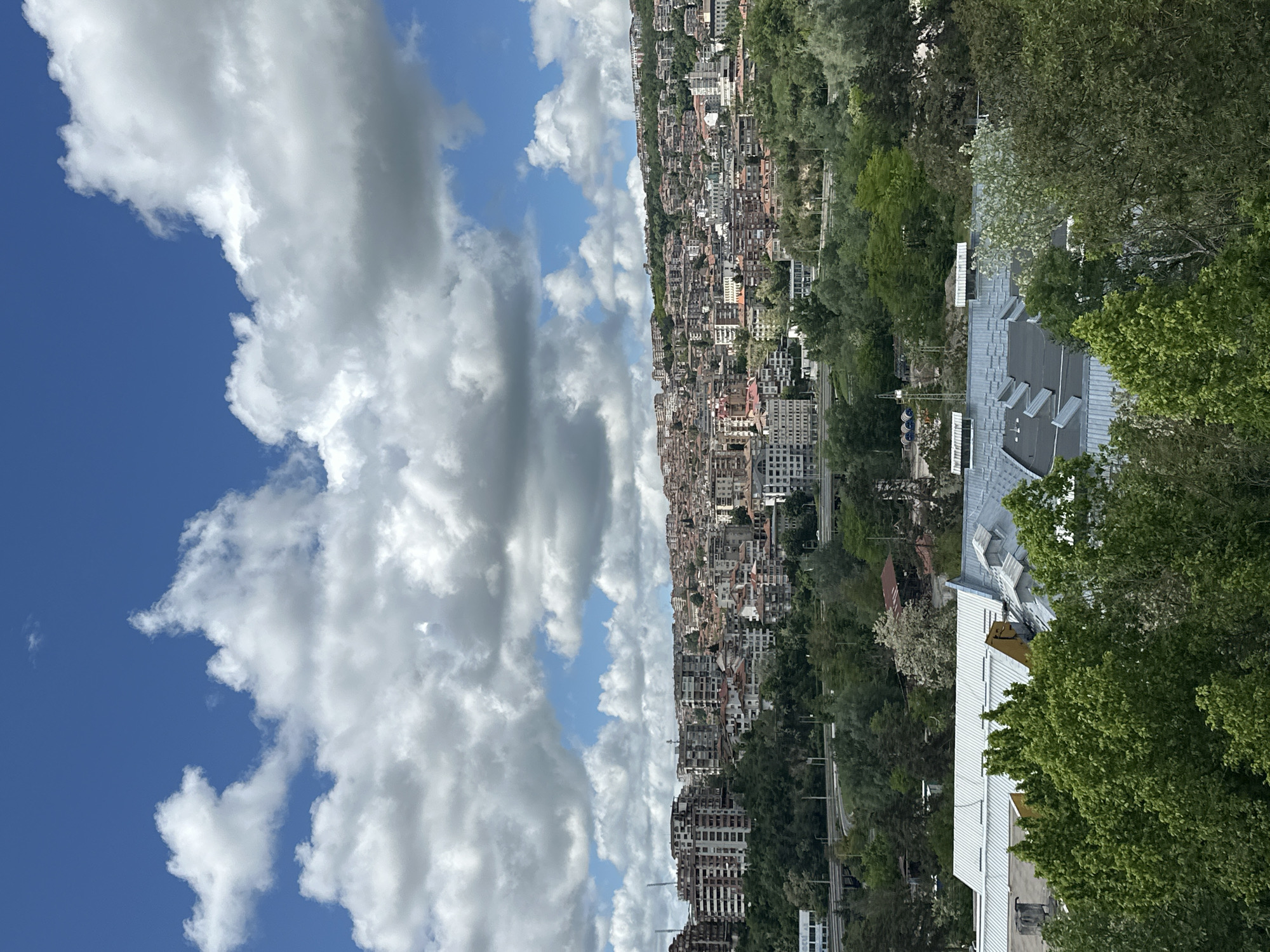 The image shows an elevated view of Veliko Tarnovo, Bulgaria, captured on a partially cloudy day. In the foreground, the corner of a white building with a gray, angled roof is partially visible, obscured by leafy green trees. The midground features a dense urban landscape comprised of buildings of varying sizes and architectural styles, intermixed with greenery. The city extends along a ridge or slope, and the buildings appear closely packed. In the background, a vibrant blue sky is filled with large, puffy white clouds. The sky occupies roughly half of the image, contrasting with the urban and natural elements below.