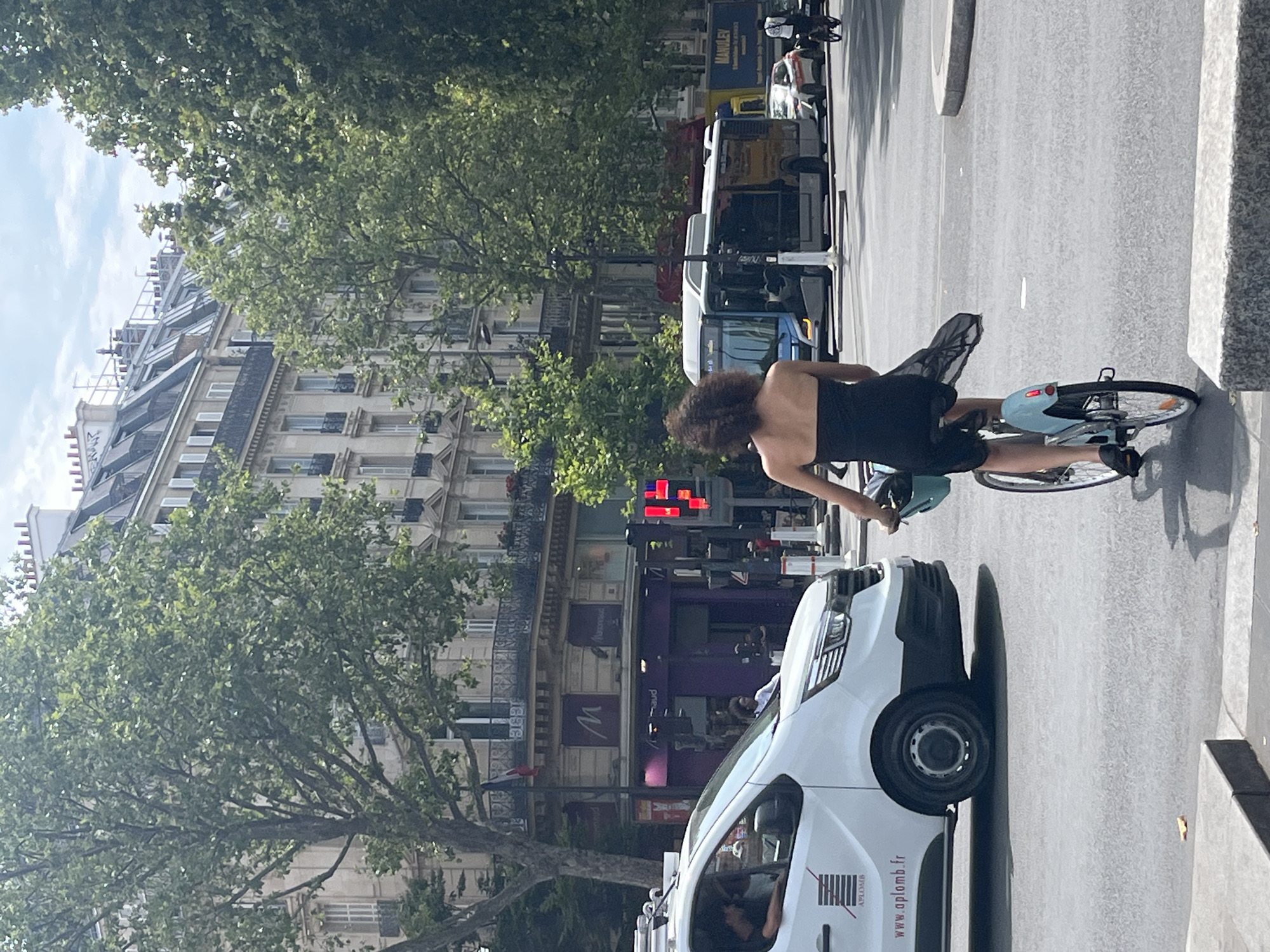 The image depicts a street scene in Paris, France, with classic Haussmannian buildings lining the background. In the foreground, a woman with dark, curly hair is riding a blue bicycle from right to left, wearing a black strapless dress. She is in the middle of an intersection, with a white van to her left. The van has a logo and website address visible. Further in the background, there are buses and other vehicles on the road, as well as traffic lights displaying red. Trees with green foliage are interspersed throughout the scene, providing shade and a natural element to the urban environment. The sky is partly cloudy, suggesting a warm day.