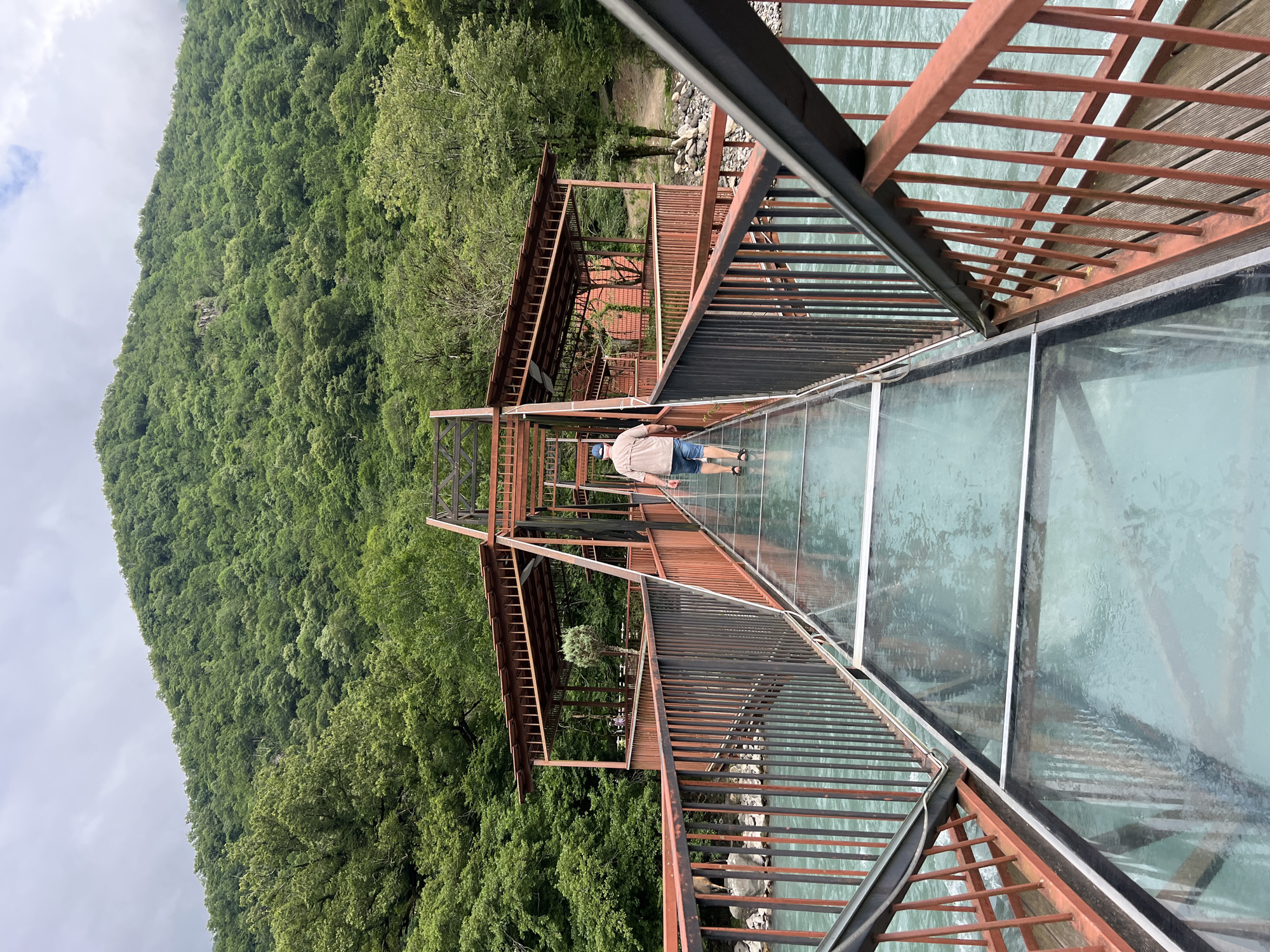 A man walks on a glass-floored bridge over a turquoise river. The bridge has red metal railings and a covered section with wooden benches. The background is dominated by a lush, green, tree-covered mountainside under a cloudy sky.