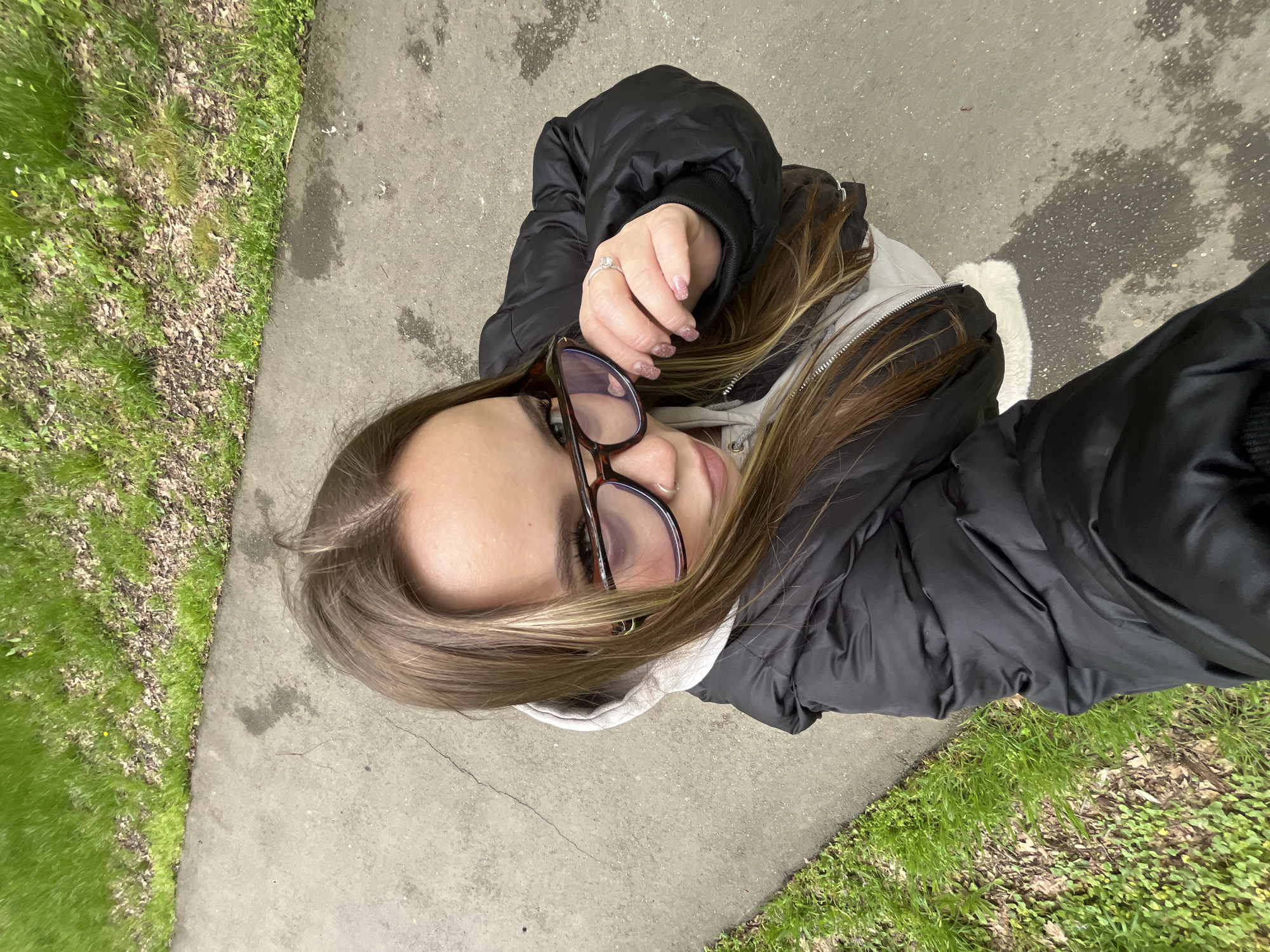 A high-angle selfie captures a young woman outdoors in Krasnodar, Russia, looking up towards the camera. The image is rotated, showing the subject from an overhead perspective, looking downwards.
The woman has long, light brown hair with blonde highlights. She is wearing dark-rimmed glasses and a small ring piercing on her left nostril. Her expression appears neutral or slightly pensive. She wears a black puffer jacket over a light-colored (possibly white or light grey) hooded sweatshirt. A silver ring with a clear stone is visible on her right ring finger, and her fingernails have a light, glittery polish.
The scene depicts an outdoor setting with a concrete or asphalt path running alongside a grassy area. The grass is green, interspersed with some brown leaves and debris, suggesting a natural, possibly urban or suburban environment. The ground surface of the path has several dark, damp patches, indicating recent moisture or stains. The overall lighting suggests daytime, possibly with overcast conditions given the lack of strong shadows. No discernible text is visible in the media.