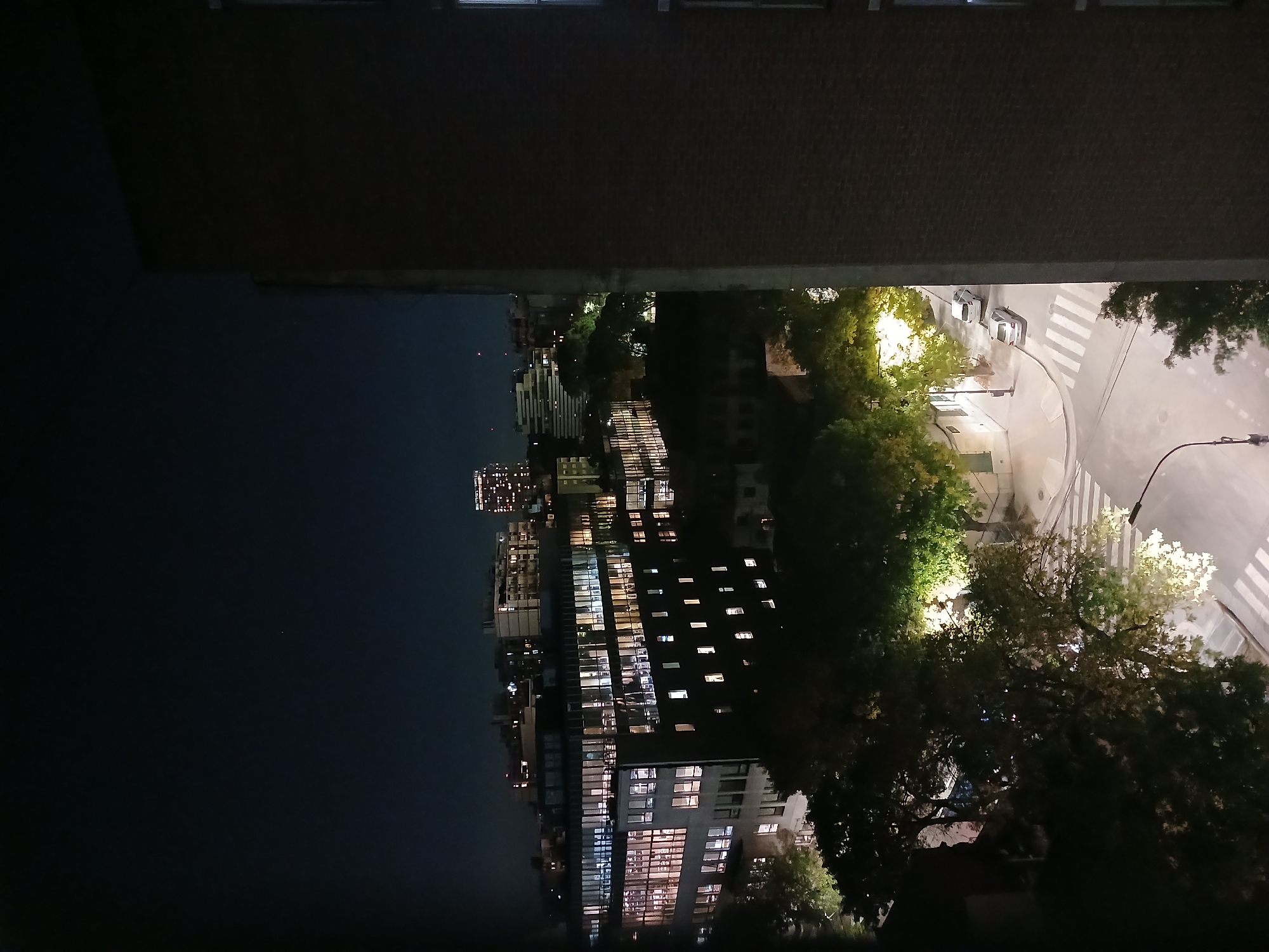 A nighttime view in Buenos Aires, Argentina, appears to be taken from a high vantage point, possibly a balcony or window. A section of a brick building frame the top portion of the image. The scene overlooks a street intersection with a crosswalk illuminated by streetlights. Several vehicles are visible on the road. Trees are present, partially obscuring the view of buildings lit up with windows at night. Further in the distance, the cityscape fades into the night sky.