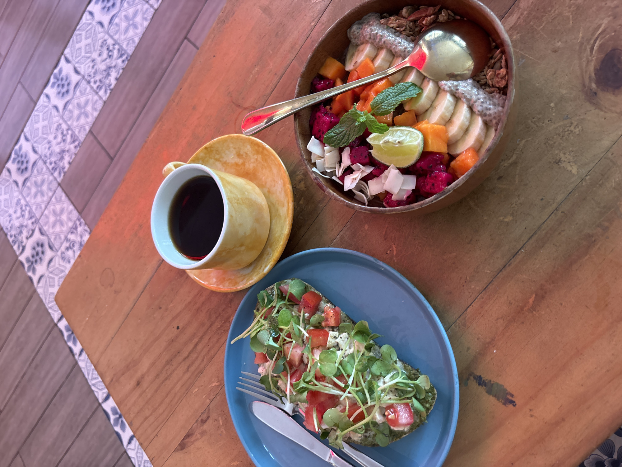 The image shows a close-up, high-angle shot of a wooden table at what appears to be a cafe in Da Nang, Vietnam, given the distinct tile flooring visible in the background with a blue and white floral pattern characteristic of Vietnamese design. On the table, there are three main food and drink items. To the left, a bright blue plate holds half of an avocado topped with diced tomatoes, crumbled feta cheese, and microgreens. A silver fork and knife are placed on the plate. To the right, a wooden bowl is filled with a colorful fruit salad, including sliced bananas, dragon fruit, papaya, mango, coconut flakes, mint leaves, and a lime wedge. A gold spoon rests within the bowl. In the upper right quadrant of the image, a small yellow ceramic mug filled with dark coffee sits on a matching yellow saucer. The wooden table has a visible grain and appears to be slightly worn. The lighting suggests ambient indoor lighting, possibly with a slight warm hue.