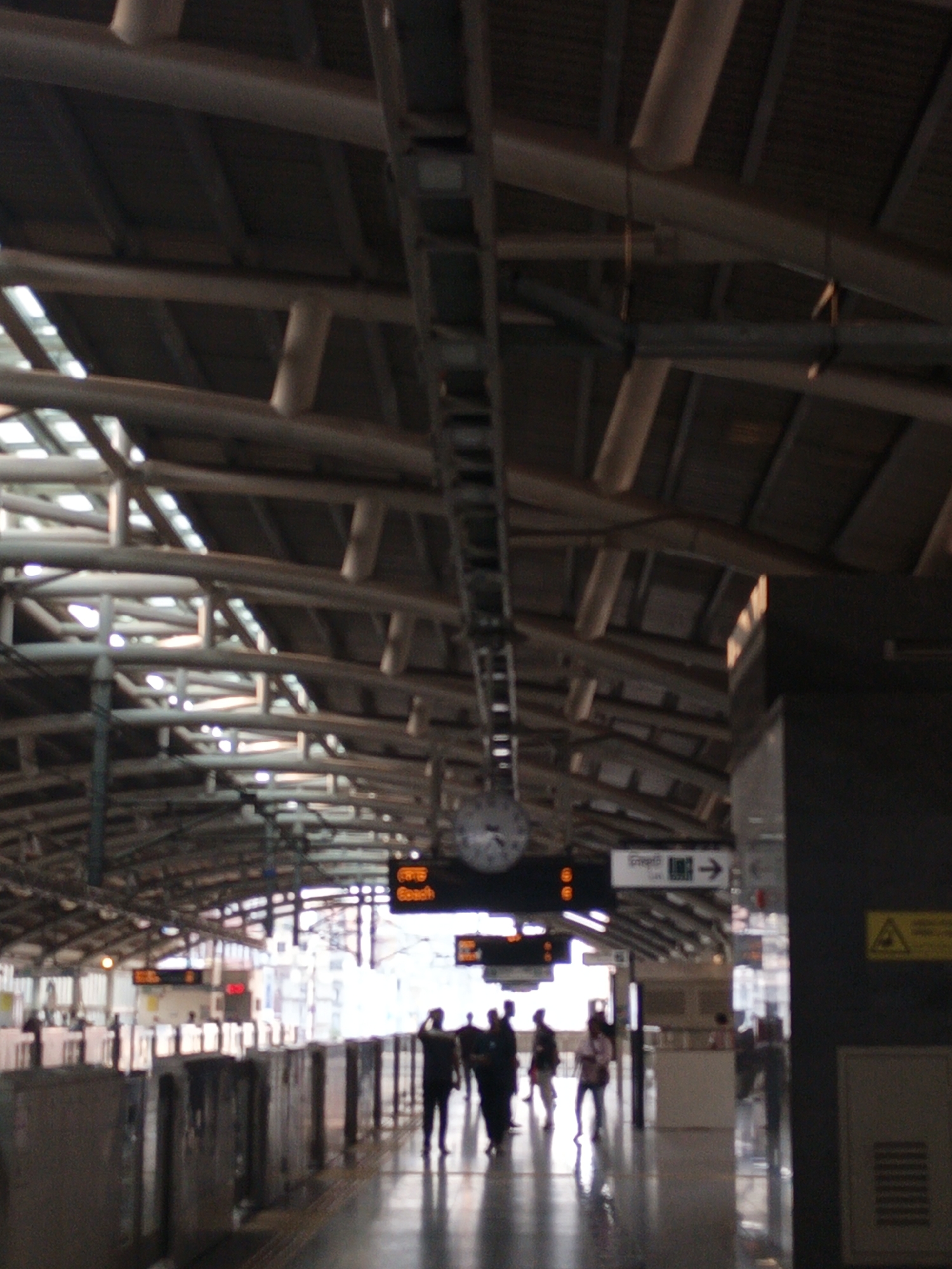 The image captures the interior of a modern metro station platform in Dhaka, Bangladesh. The scene is dominated by a complex, arched roof structure made of metallic beams and trusses, some of which appear to be wrapped or covered, with translucent panels allowing natural light to filter in.
On the platform, several people are visible, mostly as silhouetted figures due to strong backlighting coming from the far end of the station. They appear to be walking, suggesting movement typical of passengers at a transport hub. The platform itself is clean and wide, with dark flooring that reflects the ambient light and the silhouetted figures. Along the edge of the platform, there are protective barriers or gates, likely for safety.
Notable details include several digital display boards hanging from the ceiling structure. One prominent board in the mid-ground shows text in Bengali, "গোজল" (Gojol), and below it, "Beach" in English, likely indicating a destination. Another digital display shows numbers, possibly train timings or platform information. A large, circular analog clock with Roman numerals is also visible hanging from the ceiling near the center of the frame. To the right, a white sign with a black arrow points right, featuring Bengali text "প্রস্থান" (Prosthan), meaning "Exit." A yellow triangular warning sign with a black symbol is affixed to a dark pillar on the right.
The overall impression is that of a bustling yet orderly public transport environment, possibly during daylight hours given the bright light entering from the outside. The station appears well-maintained and efficiently designed for passenger flow.