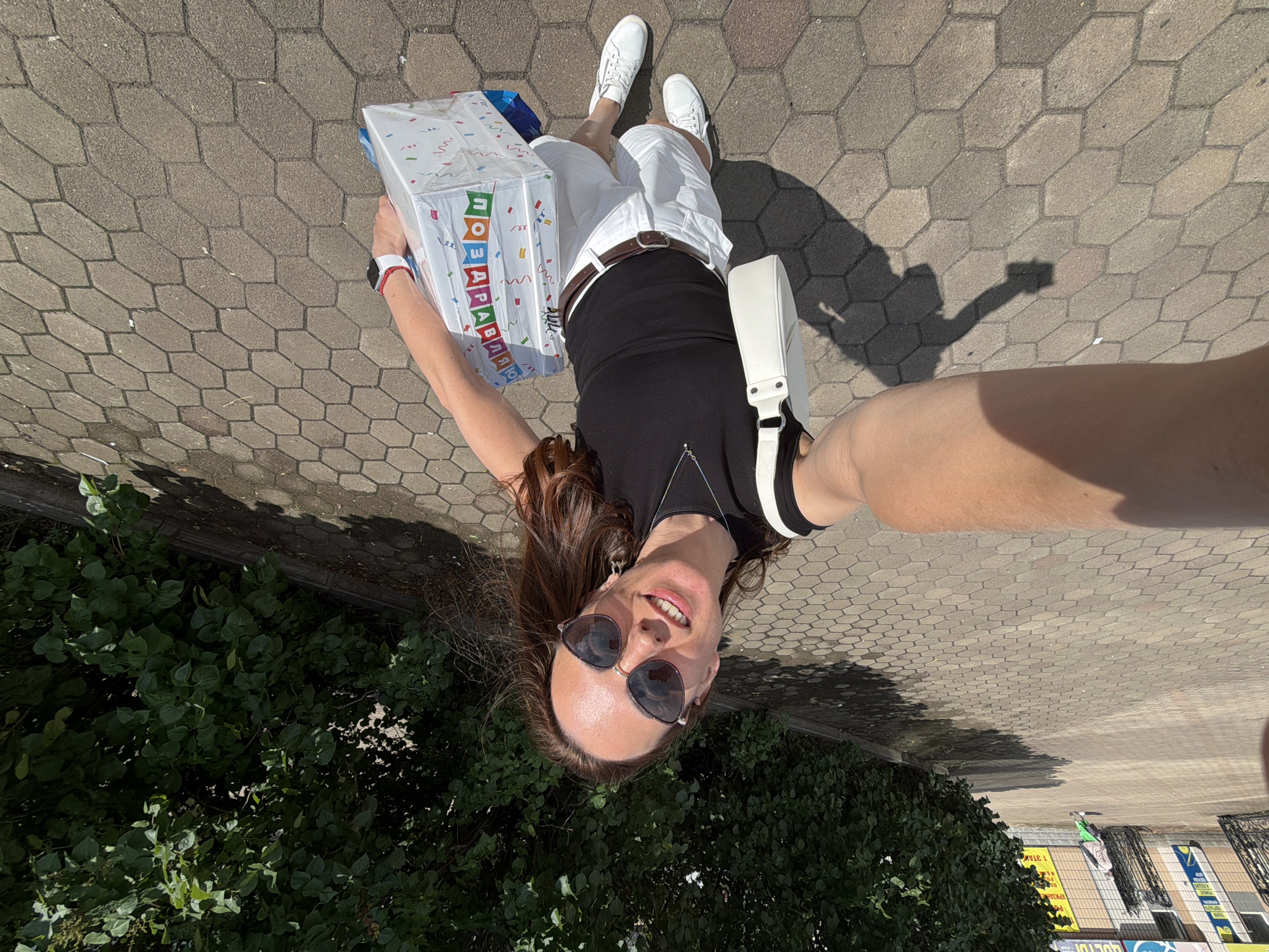 In Birobidzhan, Russia, a woman is captured in a wide-angle selfie, standing on a hexagonal-patterned paved walkway. She is facing the camera, smiling, and wearing sunglasses, a black tank top, white shorts, and white sneakers. A white purse is slung over her left shoulder, and she is holding a large, colorful gift bag in her right hand. The background features a dense green hedge and a building facade with visible signs. The sun is casting a shadow of the woman on the ground to her left. In the distance, another person wearing light-colored pants and a darker top is visible, walking away from the camera.