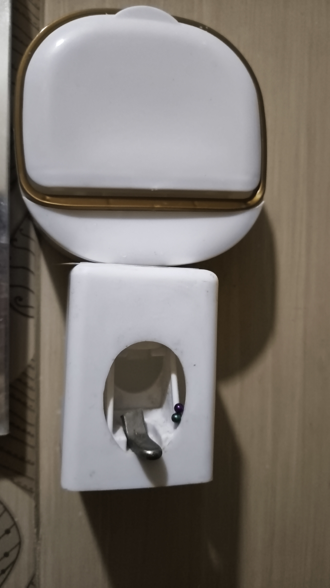The image shows a close-up view of a white plastic toothpaste dispenser mounted on a light-brown wooden wall. The dispenser has two parts: an upper rectangular section with a rounded top, outlined in gold, and a lower section with an oval opening. Inside the lower section, a silver metal lever is visible, along with three small, colorful beads (purple, blue, and green). The lighting suggests an indoor environment, likely a bathroom. The image does not provide any information about its location in South Jakarta, Indonesia.