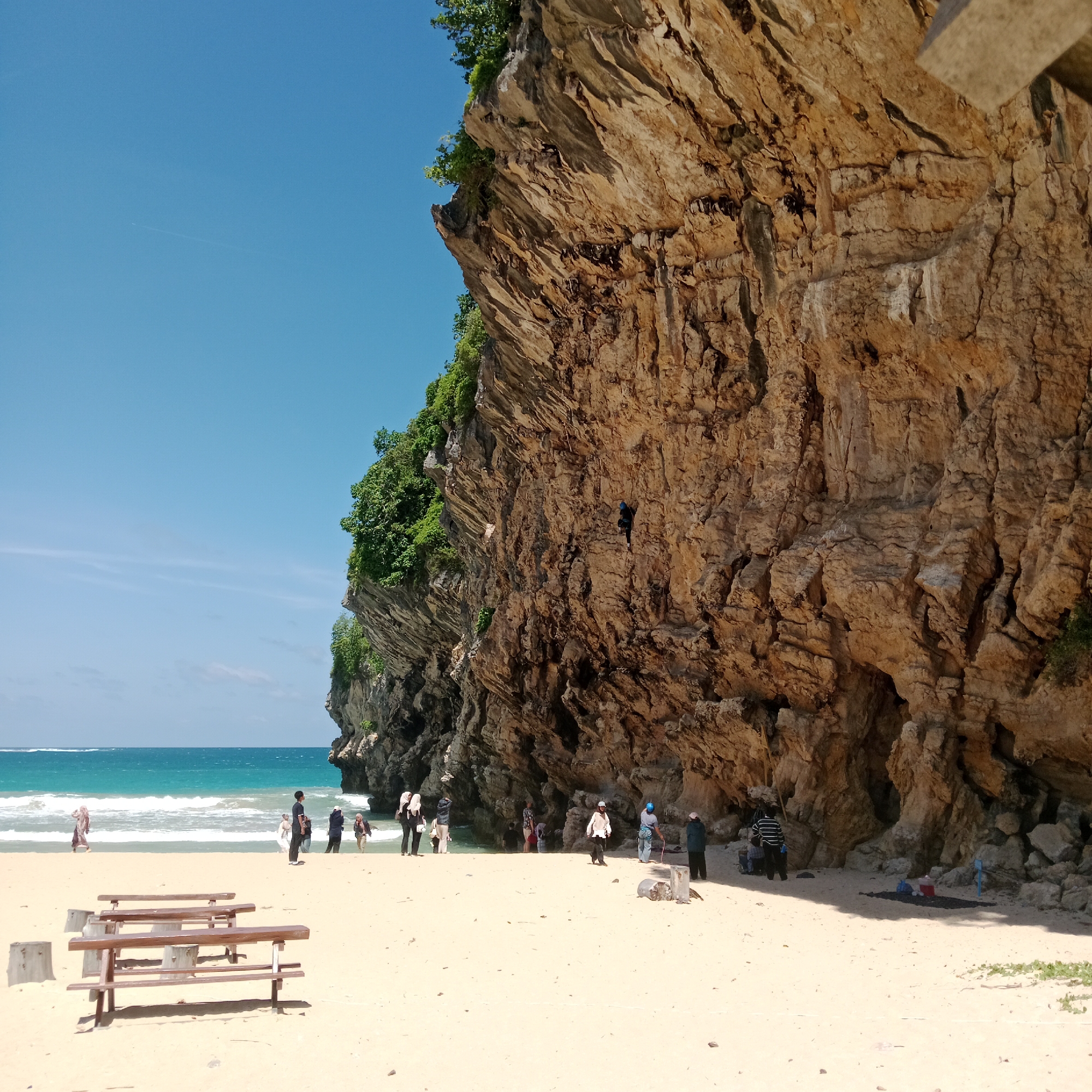 This is a medium-shot photograph taken at a beach on a sunny day, likely in Meunasahbale, Indonesia. The foreground is dominated by a sandy beach with two wooden picnic benches and two concrete block supports. In the middle ground, the ocean with turquoise water and white waves meets the sandy shore. A group of approximately ten people, dressed in casual attire and some wearing hijabs, are scattered on the beach, some walking, some standing, and a few are partially in the water. The background features a towering, textured, light brown rock cliff that curves along the coastline. A single person, dressed in dark clothing and a blue helmet, is rock climbing on the cliff face. Lush green foliage grows at the top and along parts of the cliff. The sky is a clear, bright blue.