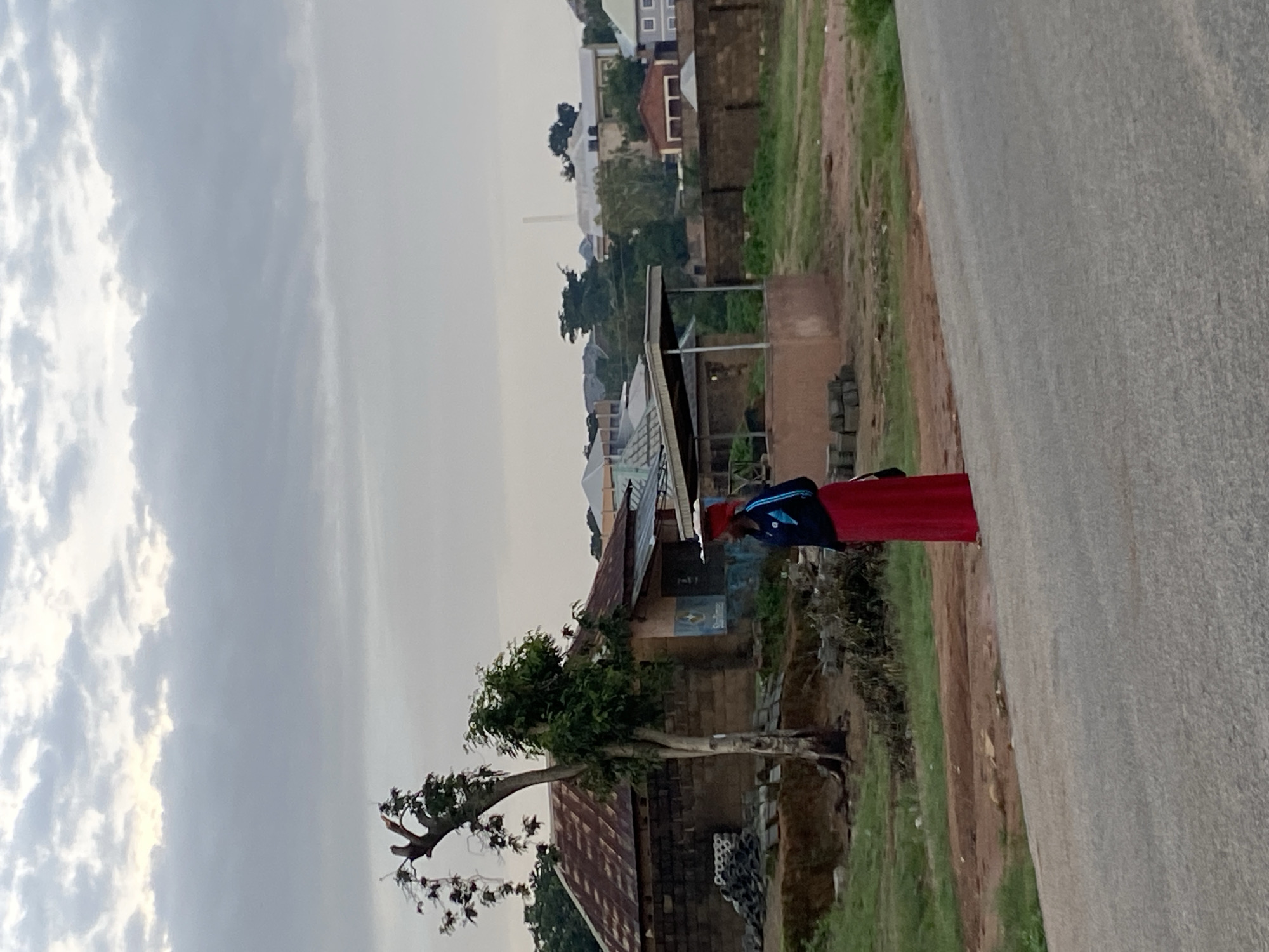 A woman in a red skirt and dark blue jacket, carrying a large flat object on her head, stands on the side of a paved road in Gwong, Nigeria. She is facing right, with a blue bag slung over her shoulder. Behind her, a small, rustic building with a corrugated metal roof is visible, as well as a lone, thin tree with sparse foliage. The background shows more buildings and lush green vegetation under a cloudy sky. The environment suggests a rural or semi-urban setting.