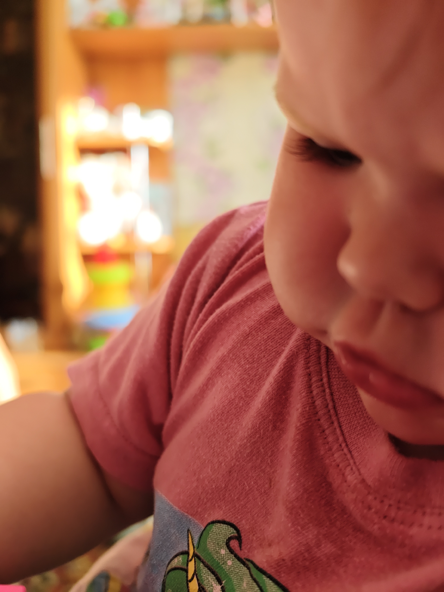 A close-up shot focuses on the face and upper body of a young child wearing a pink t-shirt. The child is looking down, with their nose and lips in clear view. The background is blurry, showing what appears to be a shelf or cabinet. There is nothing to suggest the picture was taken in Уральское, Russia.