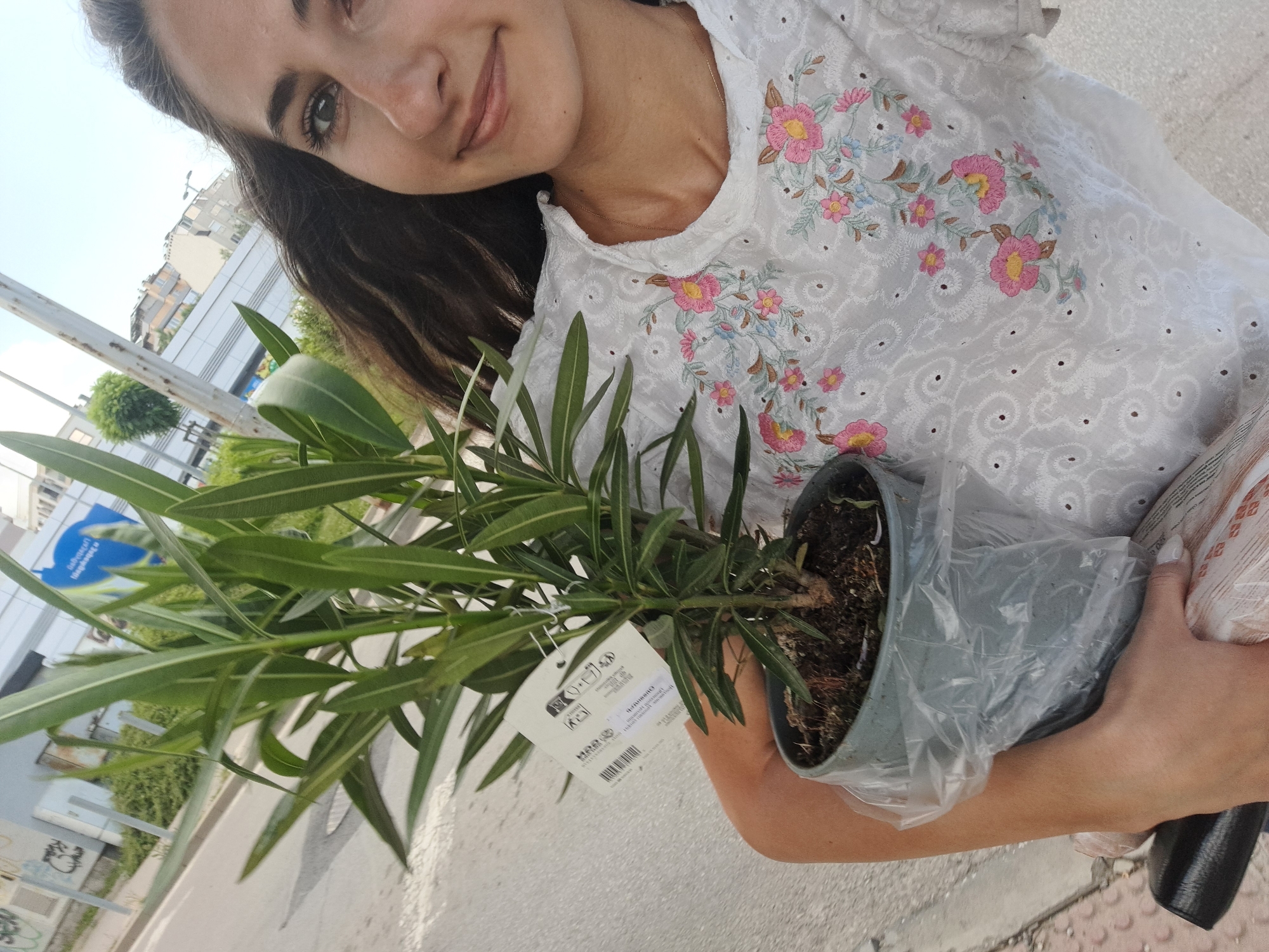 A young woman with dark, curly hair is holding a potted plant in her left arm. She is wearing a white, embroidered top with pink and blue floral patterns. The plant has long, green leaves and is in a grey pot wrapped in plastic. She is outdoors in what appears to be a city environment, with buildings and a street visible in the background. The lighting suggests it is daytime.