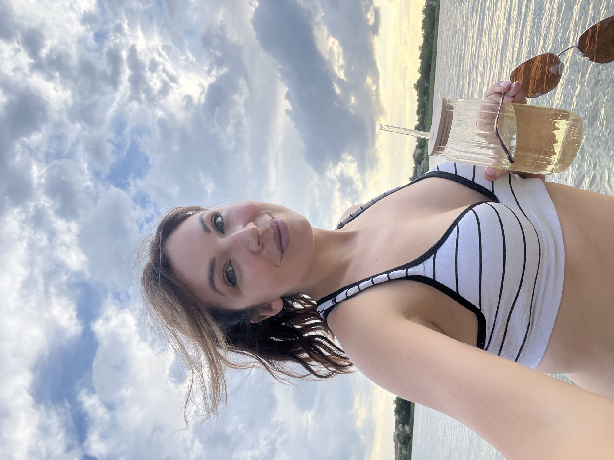 A woman is taking a selfie in a striped bikini, holding a glass of yellow liquid and sunglasses. She is standing in front of a body of water with a cloudy sky. The background shows a shoreline with houses and trees. The location is Lozuvatka, Ukraine.