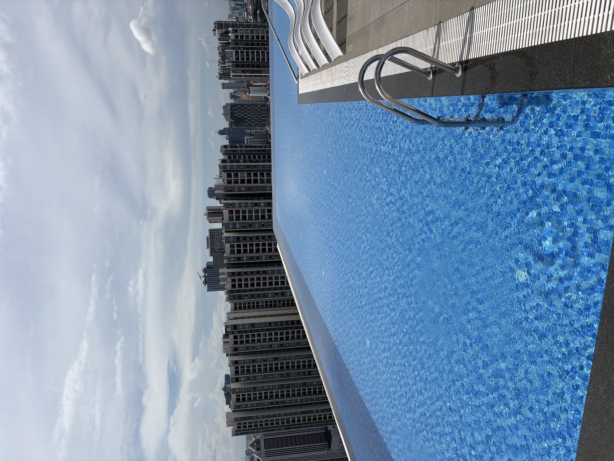 This image depicts a rooftop infinity pool overlooking a dense urban skyline in the city of Khan Mean Chey, Cambodia. The pool is a vibrant blue, tiled with small, square mosaic tiles, and features a stainless steel ladder for entry. The pool's edge appears to be lined with dark granite. To the right of the pool, several modern, white lounge chairs are arranged on a gray tiled deck, which is bordered by a white drainage grate.
In the background, a vast expanse of buildings stretches across the horizon, characteristic of a major city. The architecture is predominantly modern, with numerous high-rise apartment buildings and office towers. The buildings are a mix of gray, white, and darker tones, with repeating window patterns indicating many floors. The skyline suggests a densely populated and developing urban center.
The sky above is partly cloudy, with large, diffused white clouds against a pale blue background, indicating it is daytime. The lighting suggests it might be overcast or late afternoon, as there are no harsh shadows. The scene conveys a sense of luxury and urban living, with the pool offering a recreational amenity amidst the bustling city. No people are visible in the image.