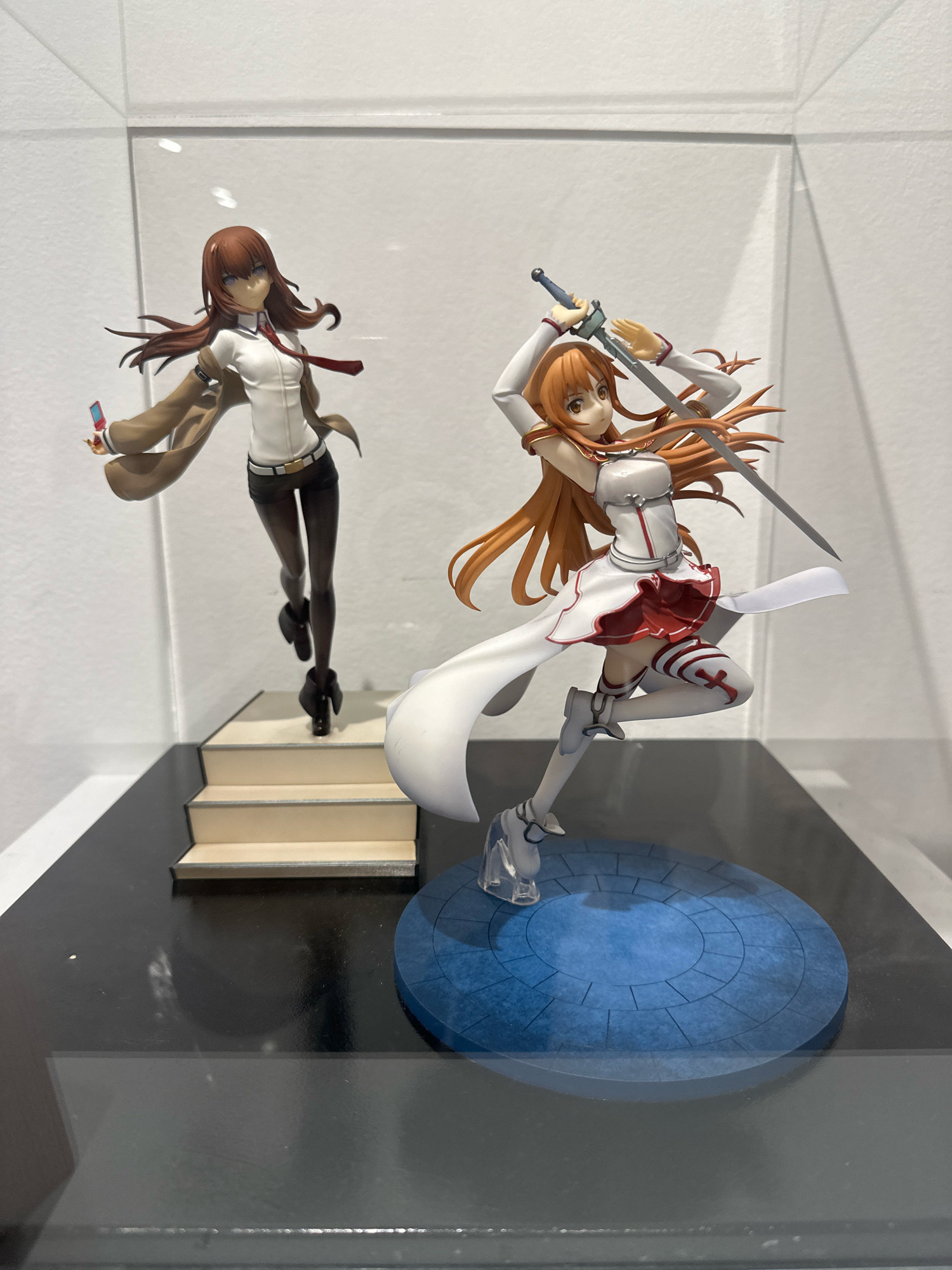 Two anime figurines are displayed in a glass case. The figurine on the left depicts a woman with brown hair, wearing a white shirt, red tie, brown coat, and black shorts, holding a pink device. She is standing on a set of tan stairs. The figurine on the right shows a woman with long orange hair, wearing a white and red outfit and white boots, holding a sword. She is posed in a dynamic stance on a blue circular base with a stone pattern. The background is plain white, and the figurines are placed on a black surface. There is no indication of the city of Belgrade, Serbia, or any other specific location.