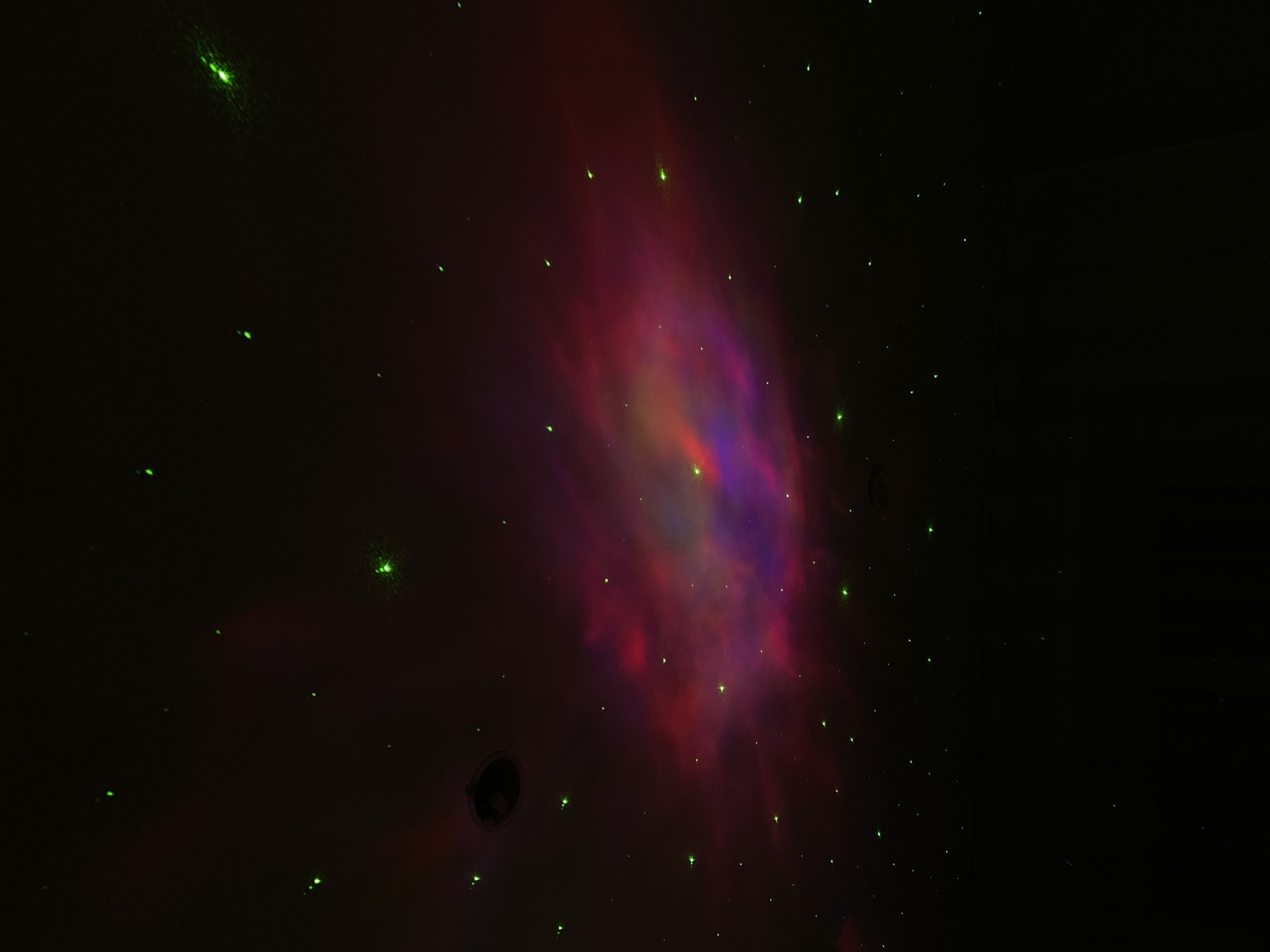 The image shows a dark, indoor space, possibly a room, with a red and purple nebula-like effect projected on a surface, likely a ceiling. Scattered across the scene are numerous small, green laser-like dots. There's a dark circular object that appears to be a vent or speaker on the surface. The background is mostly black. There is nothing in the image that indicates a location in Votkinsk, Russia.