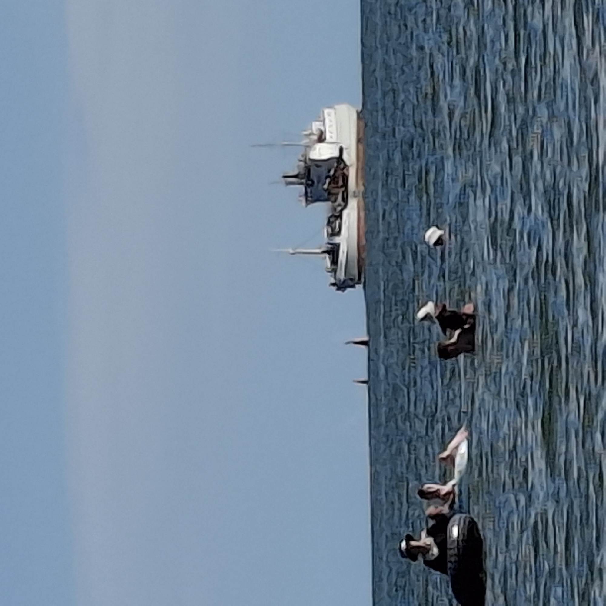 In the open water under a clear blue sky, a group of people are enjoying leisure activities. A large white and grey ship is visible in the distance, on the horizon. In the foreground, several individuals are in the water. One person, wearing a hat, is on a dark inflatable tube. Another person is partially submerged in the water, with an arm raised. Further to the right, two more people are floating, one with a white object on their head. The water is a deep blue with visible ripples, suggesting a calm day. The scene conveys a sense of relaxation and outdoor recreation.