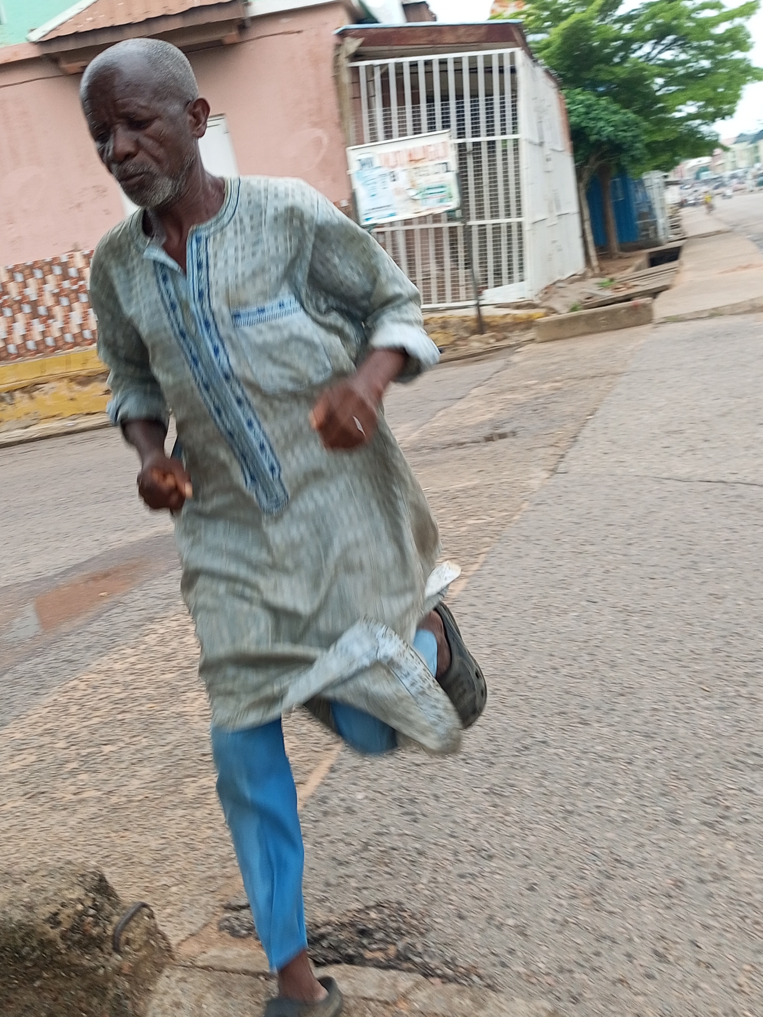 The image shows an elderly African man with a shaved head and a grey beard running on a paved street in Arawa, Nigeria. He is wearing a light grey kaftan with blue trim and bright blue trousers. His left leg is mid-stride, and his arms are bent at the elbows, indicating forward motion. The man appears to be alone. The background shows buildings and a street with some vegetation. The overall scene is that of a man exercising outdoors in an urban setting.